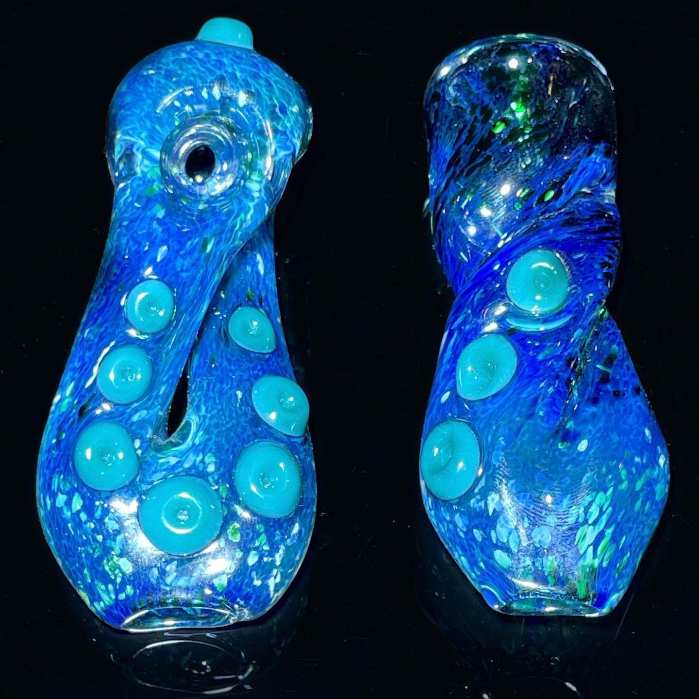 Two blue hand-blown glass sculptures with embedded bubbles, illuminated from within, set against a black background.
