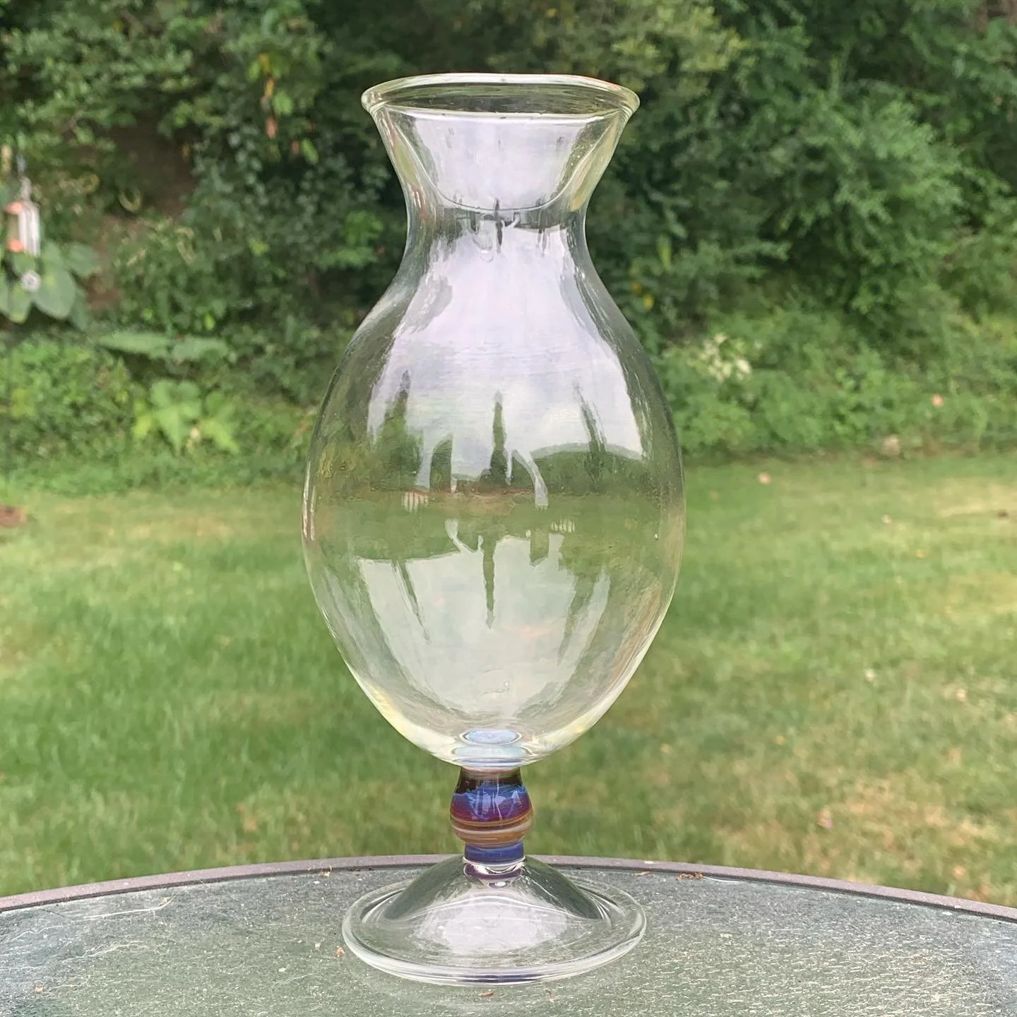 A clear glass vase with a wide top, a narrow middle, and a rounded base, sitting on an outdoor surface with grass and trees in the background.