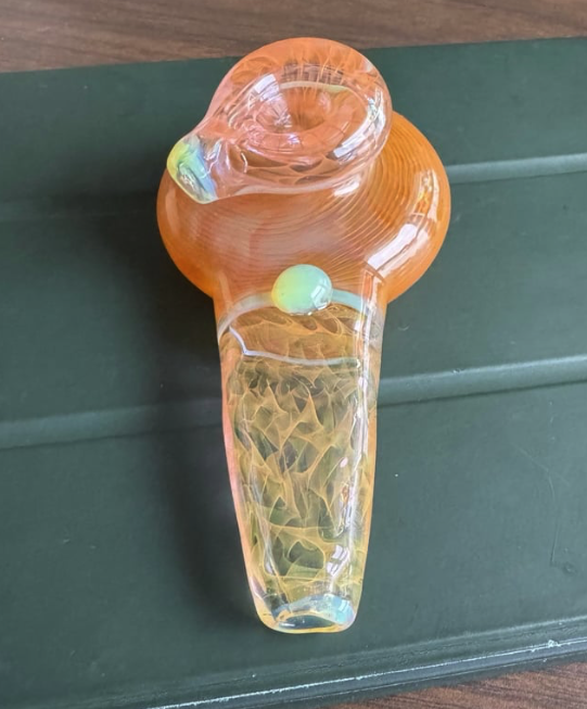 A glass smoking pipe with orange and yellow hues resting on a dark green surface.