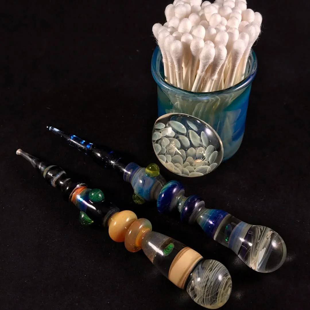 Glass smoking pipes with colorful patterns, a container of cotton swabs, and a small mirror on a black surface.
