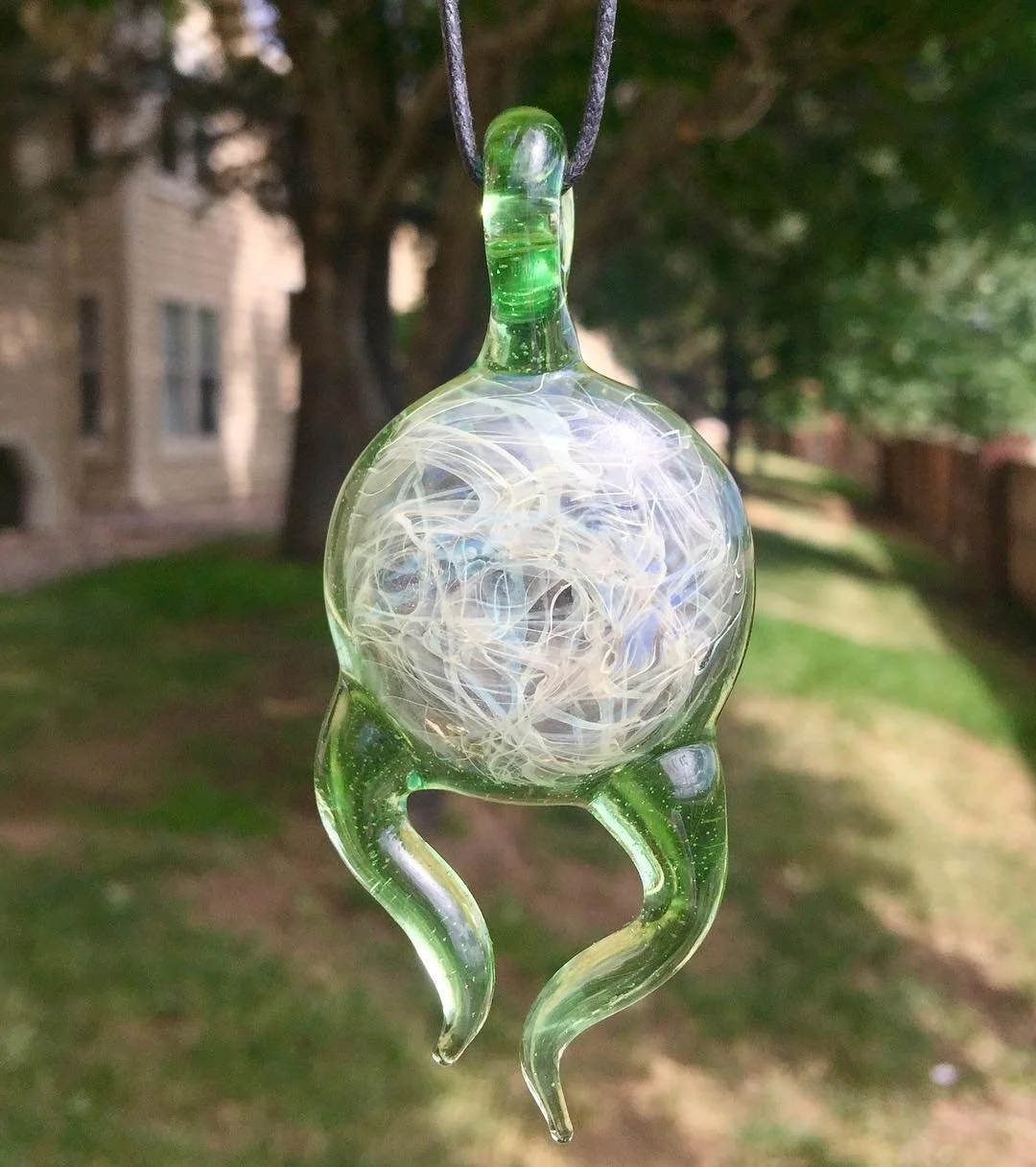 Glass ornament with a white, swirling design inside, hanging outdoors with trees and a brick building in the background.