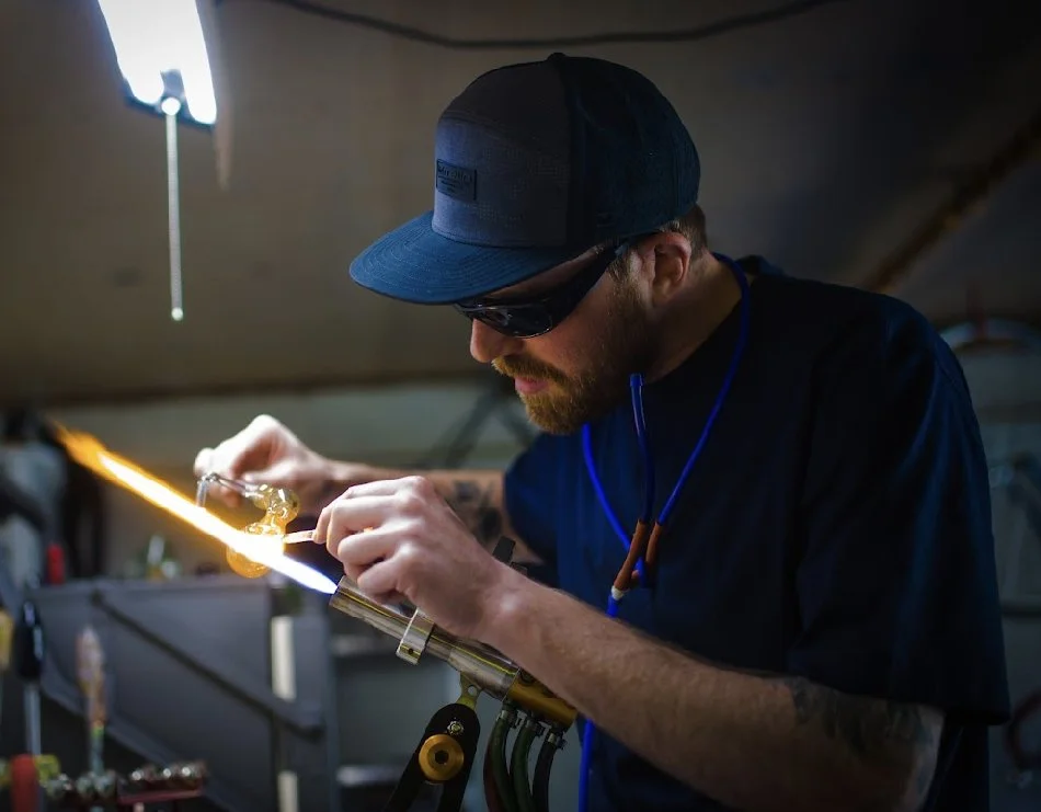 A man working on glassblowing, using a torch torch to melt and shape glass in a workshop.