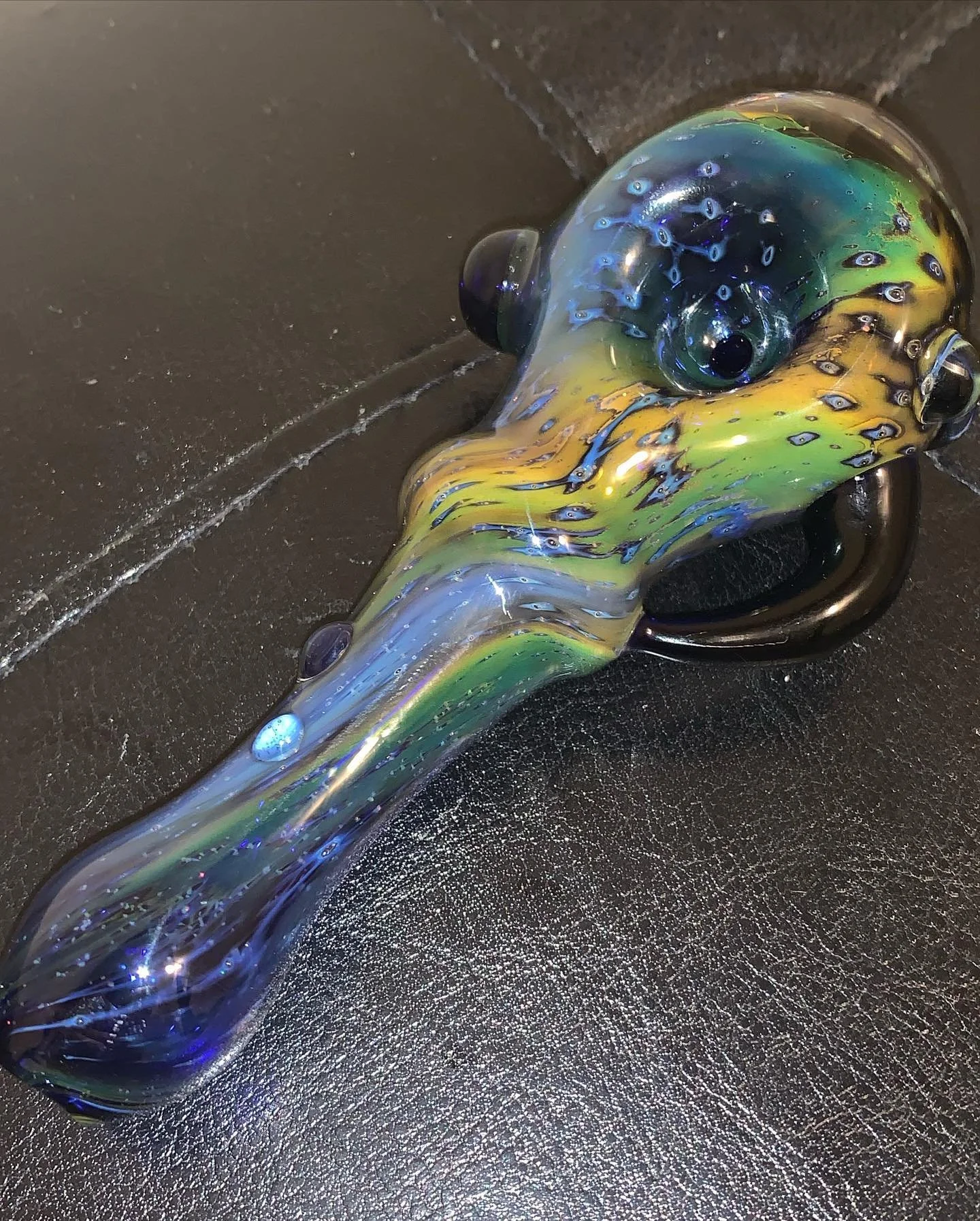 A colorful glass pipe with a design resembling a fish, featuring vibrant shades of green, yellow, blue, and black, resting on a dark textured surface.
