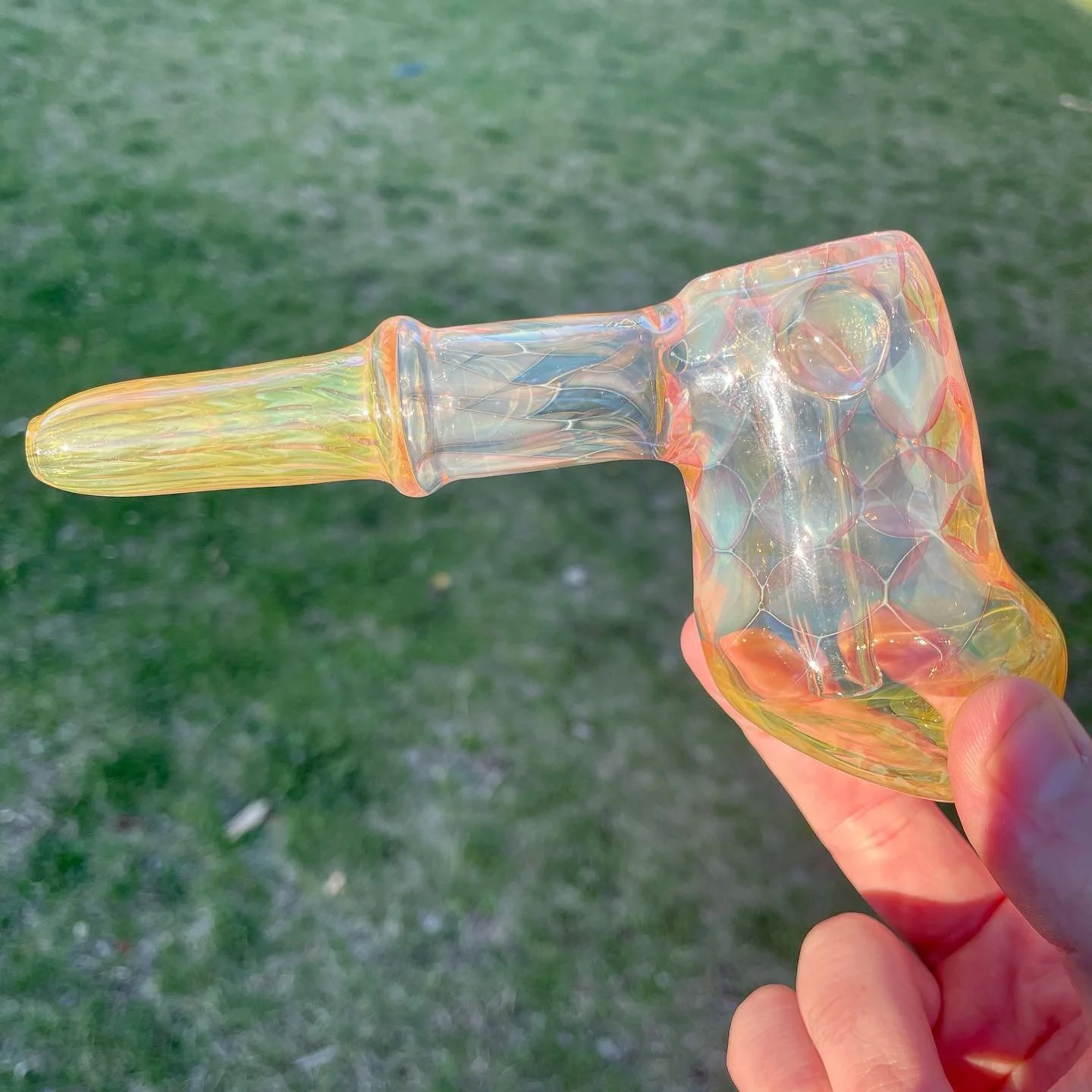 A person holding a colorful glass pipe, designed in the shape of a unicorn, against a grassy outdoor background.