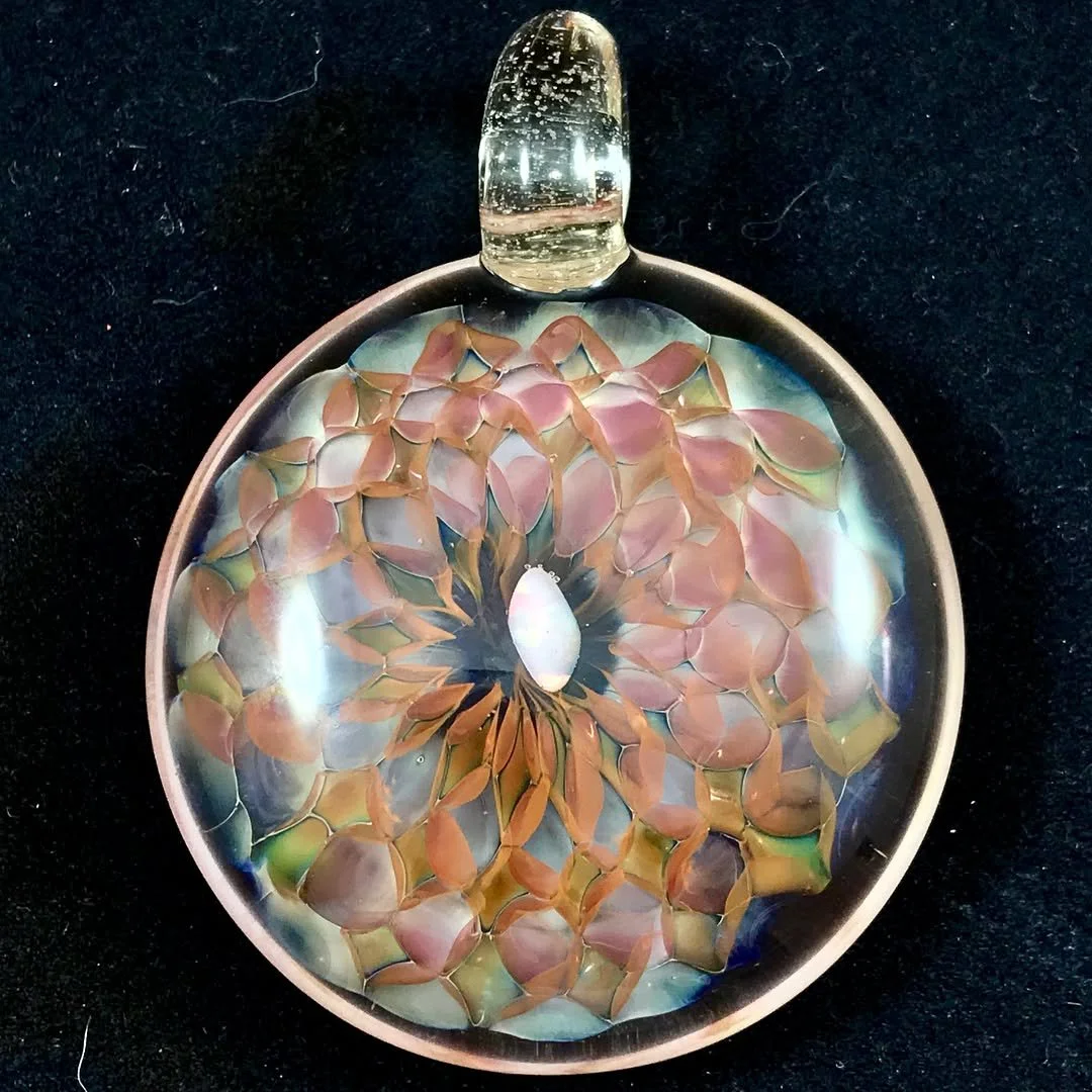 A glass art pendant with a gold-tone outline, featuring an abstract design with pink, blue, green, and black colors, placed on a black surface.