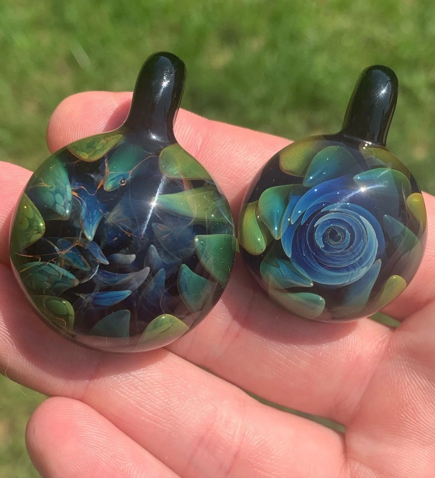 Two glass pendants with blue and green floral patterns, held between fingers outdoors in sunlight.