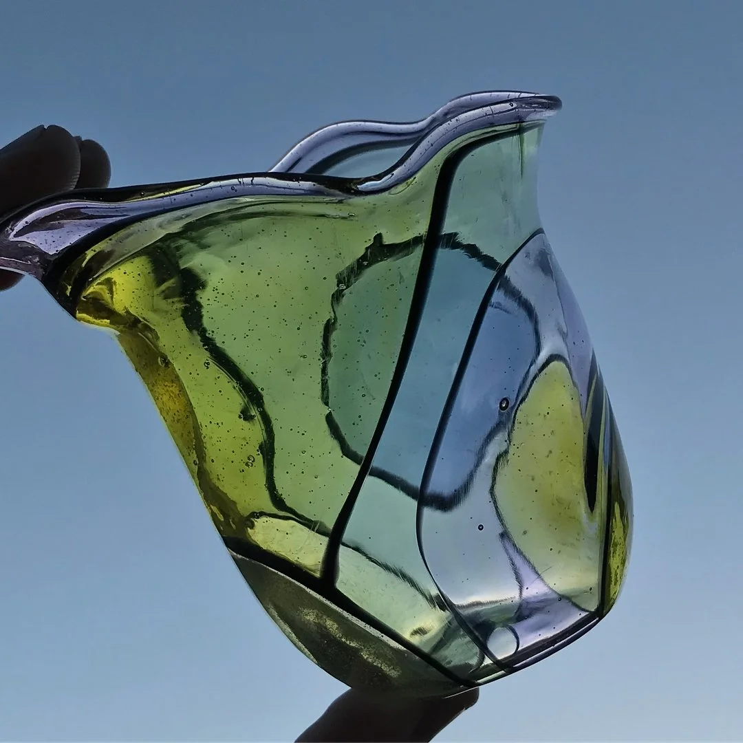 Close-up of a hand holding a piece of colorful, abstract glass art against a clear sky background.
