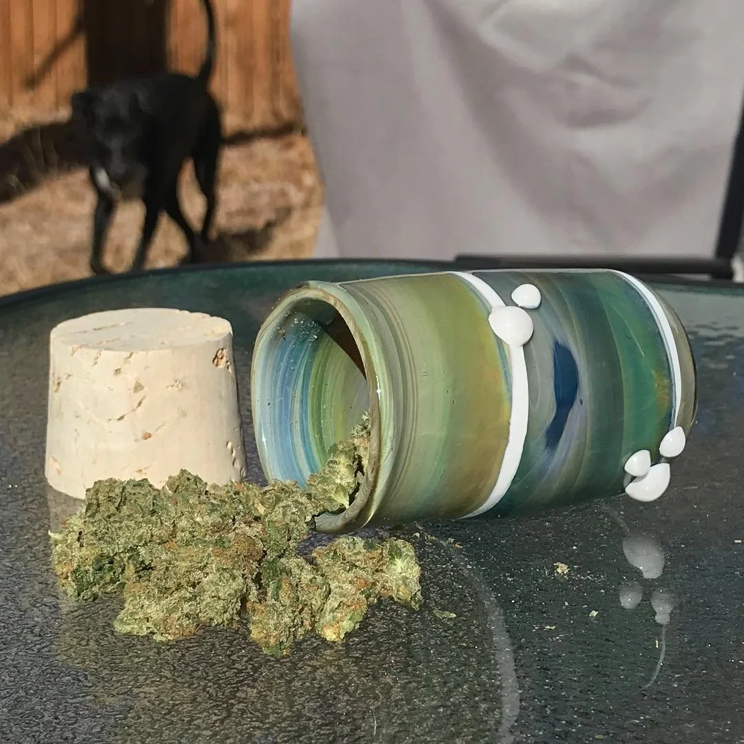 An overturned multicolored marble jar with white spots, a few cannabis buds, and a cork on a dark surface with a black dog in the background.