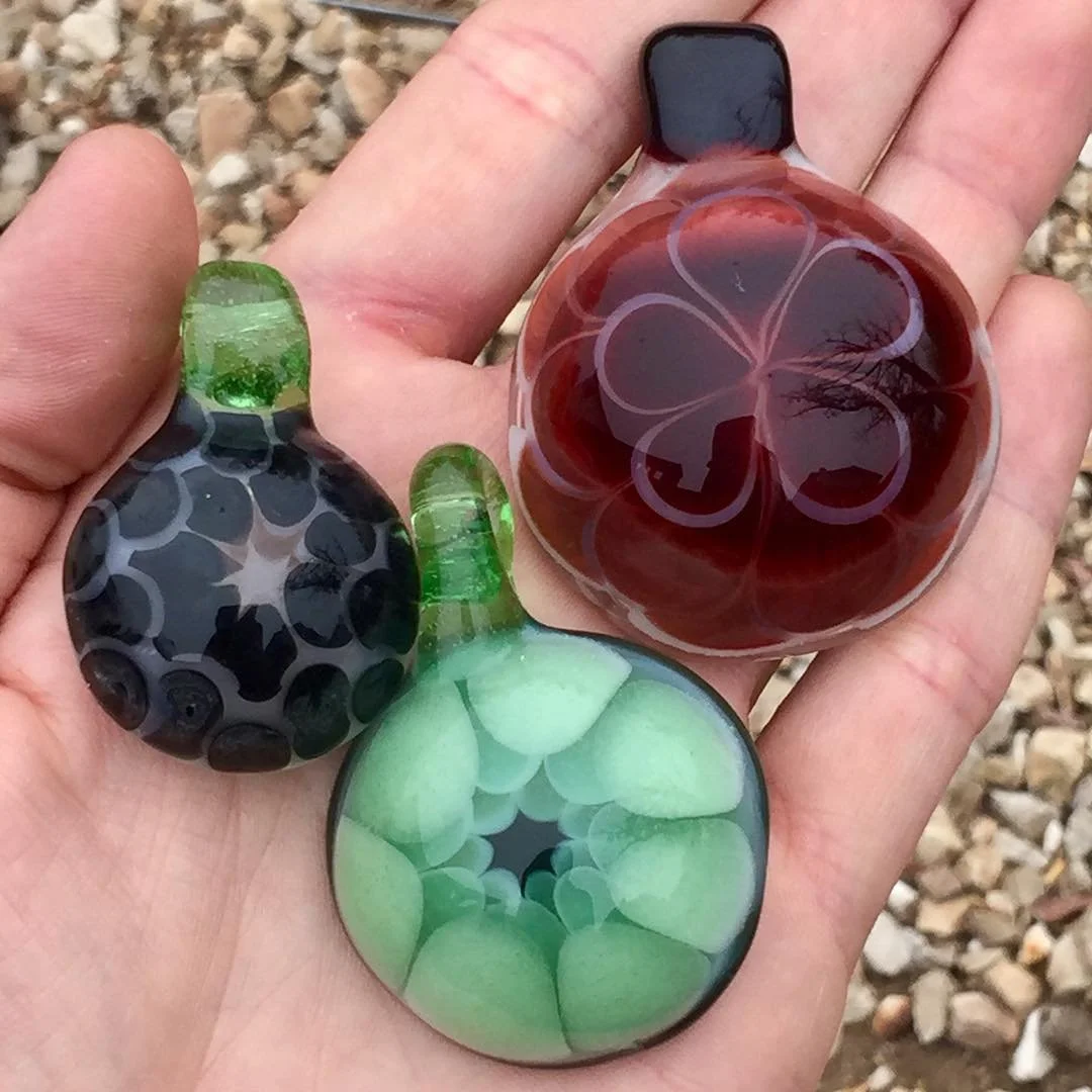 Hand holding three colorful glass paperweights outdoors with rocky ground in background.