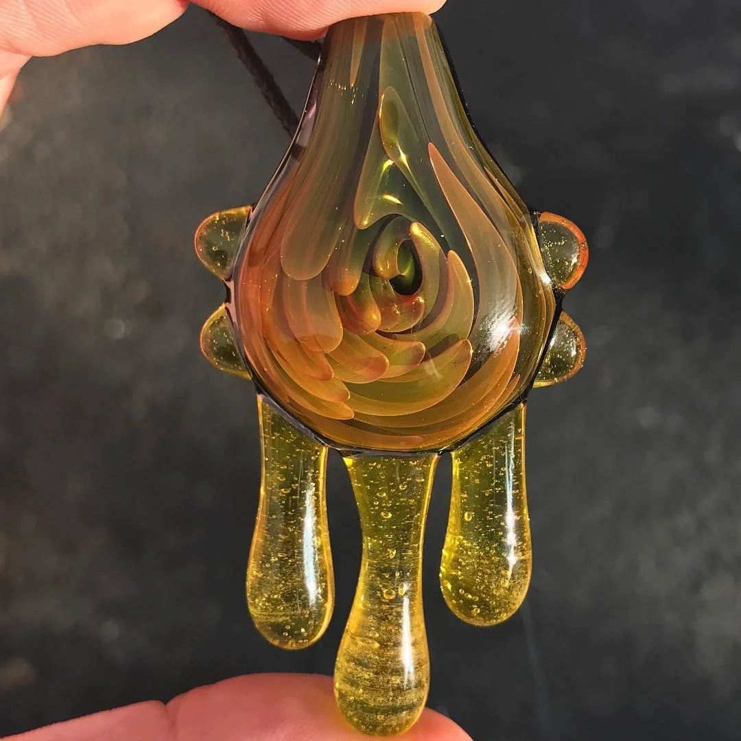 Hand holding a glass art piece resembling an abstract droplet with swirling colors and elongated hanging glass droplets.