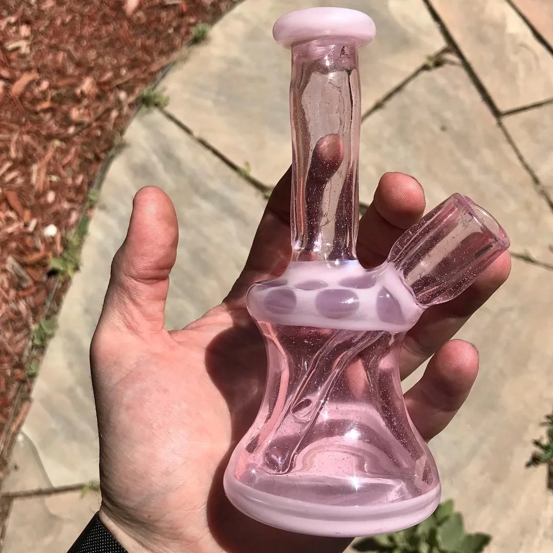 Hand holding a small pink glass bong with a curved design outdoors on a stone pathway with red mulch and greenery.