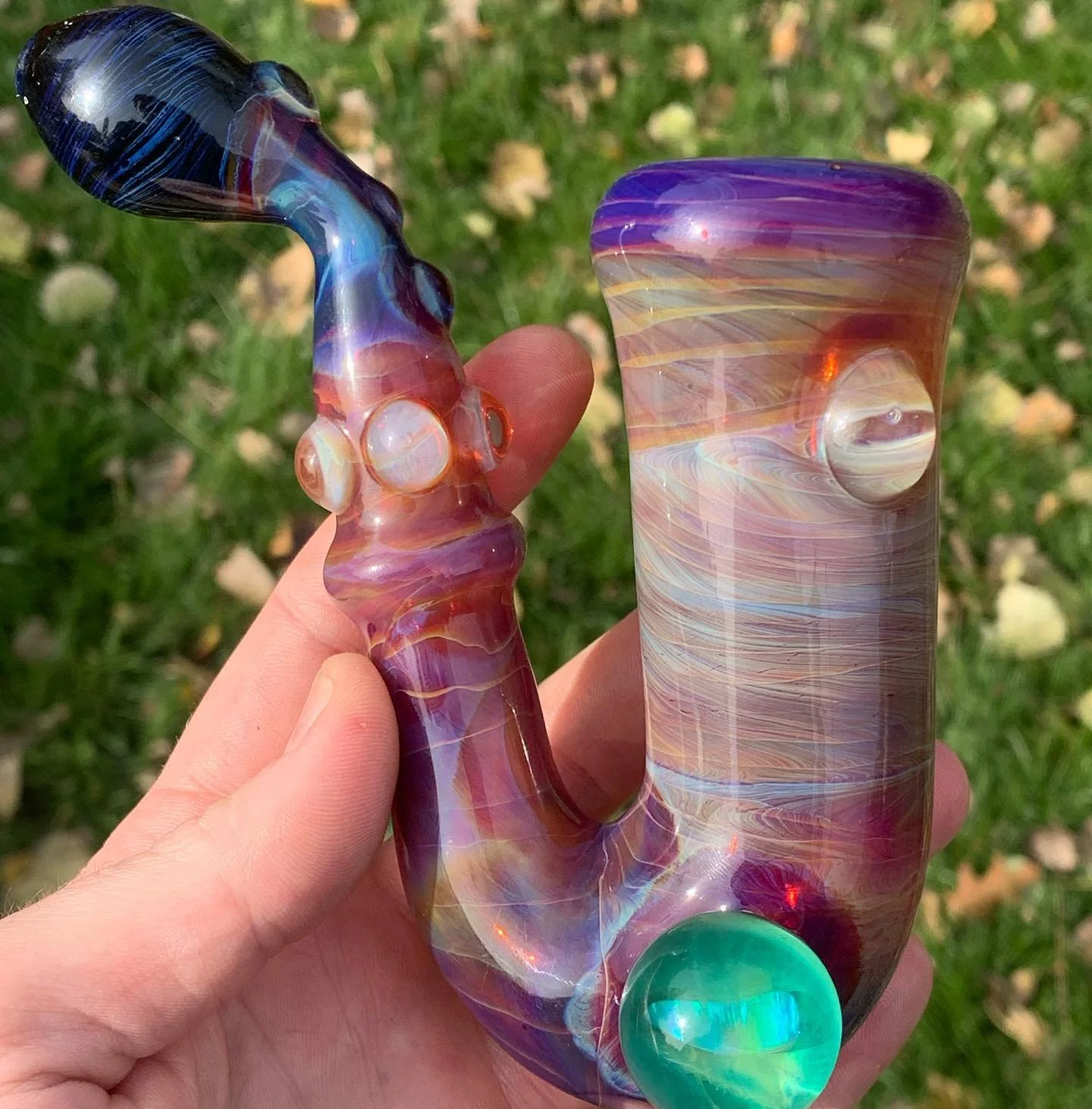 Colorful glass pipe with swirled pink, orange, purple, and blue patterns held by a person's hand outdoors on grass.