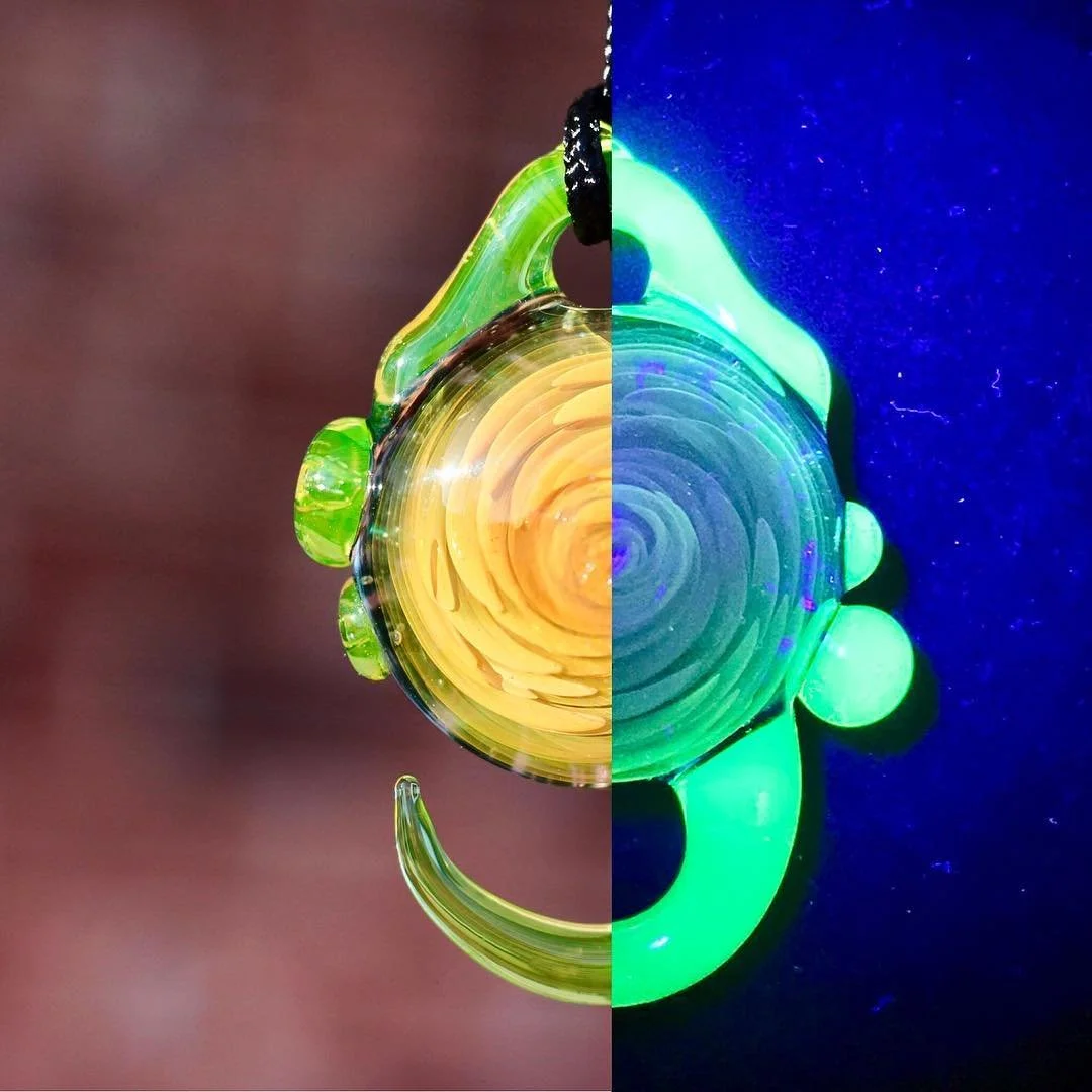 Close-up of a glass pendant pendant with green and yellow swirling colors, half illuminated in natural light and half glowing under UV light with a dark blue background.