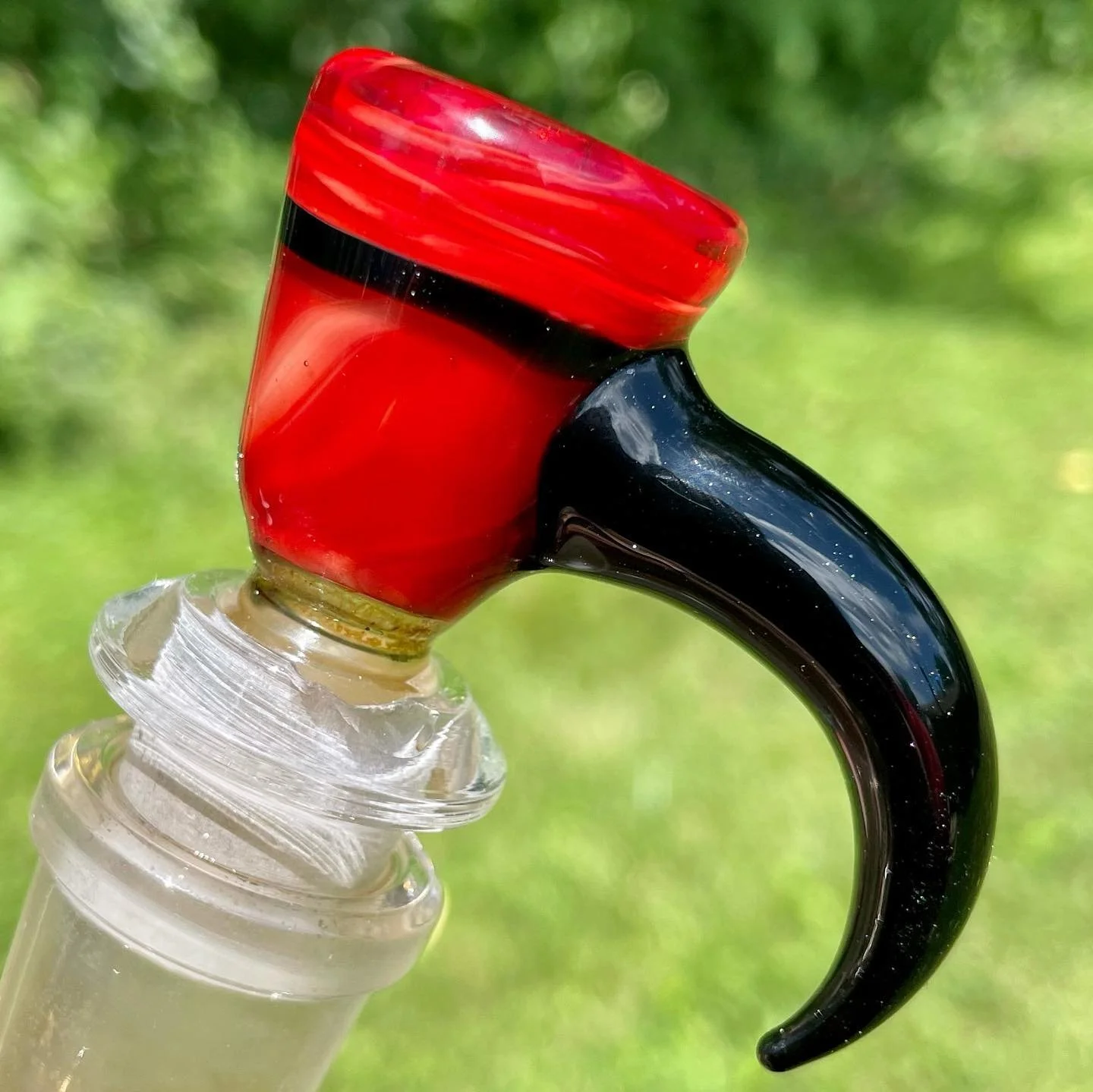 A close-up of a glass pipe with a black mouthpiece, red bowl, and clear bulb, set against a blurred green background.