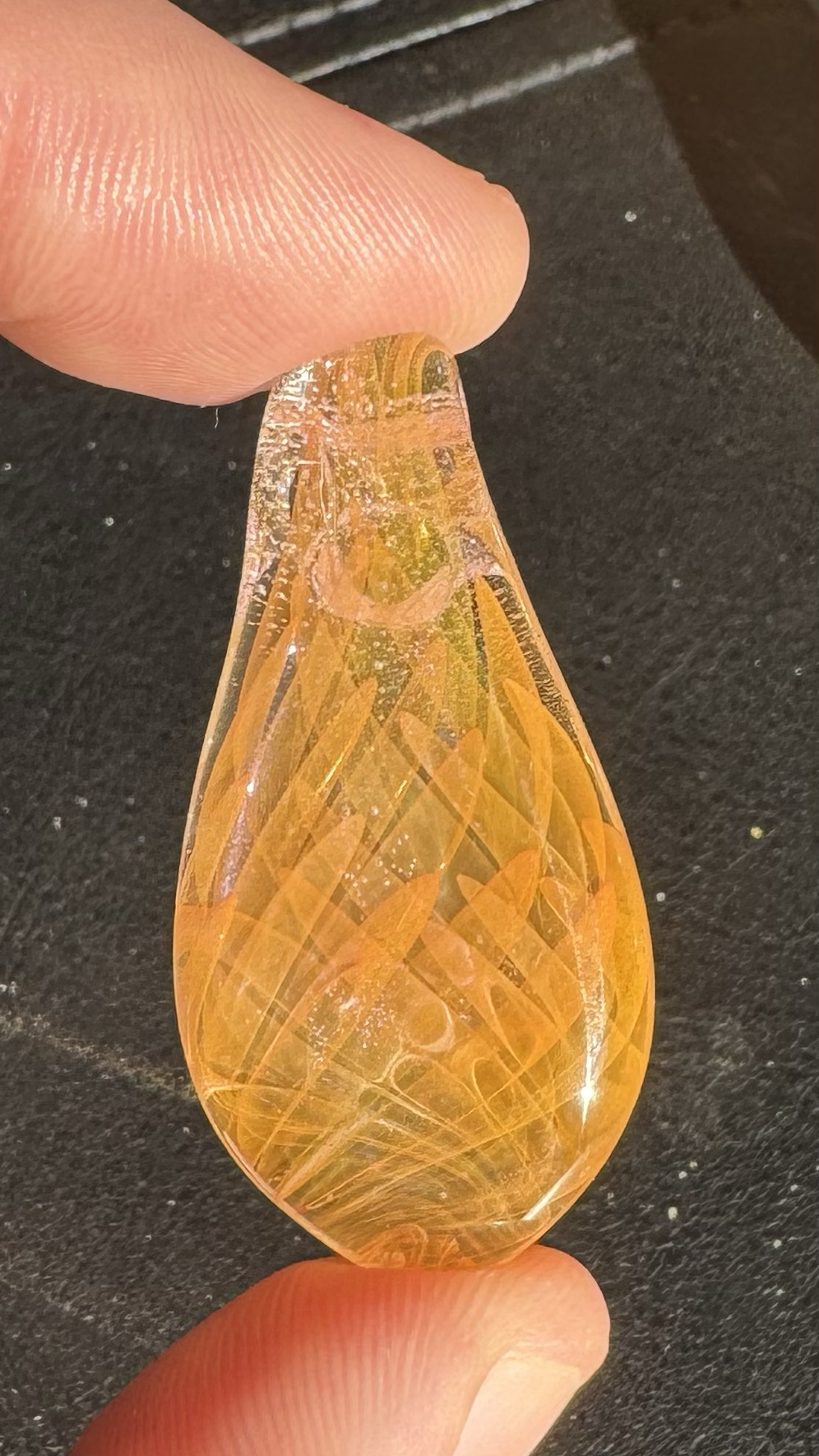 A close-up of a hand holding a polished, teardrop-shaped, orange gemstone with internal patterns.