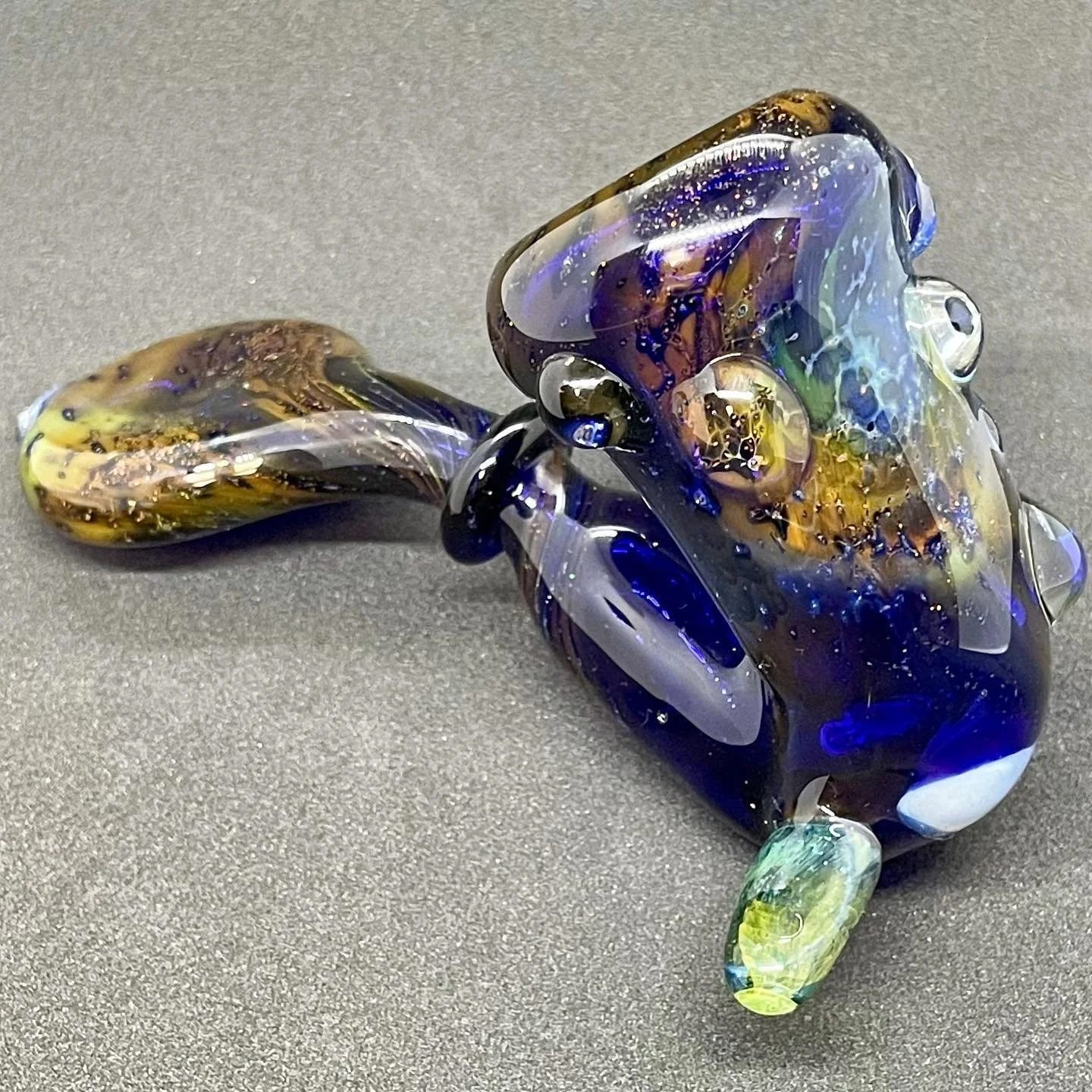 Colorful glass art sculpture resembling an abstract animal figure with blue, yellow, and brown hues, lying on a gray surface.
