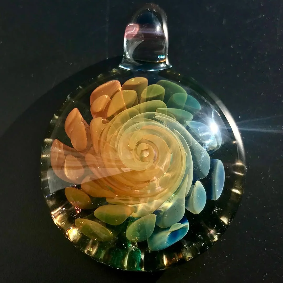 A glass pendant with a colorful spiral pattern resembling a rose, featuring shades of yellow, orange, green, and blue, set against a black background.