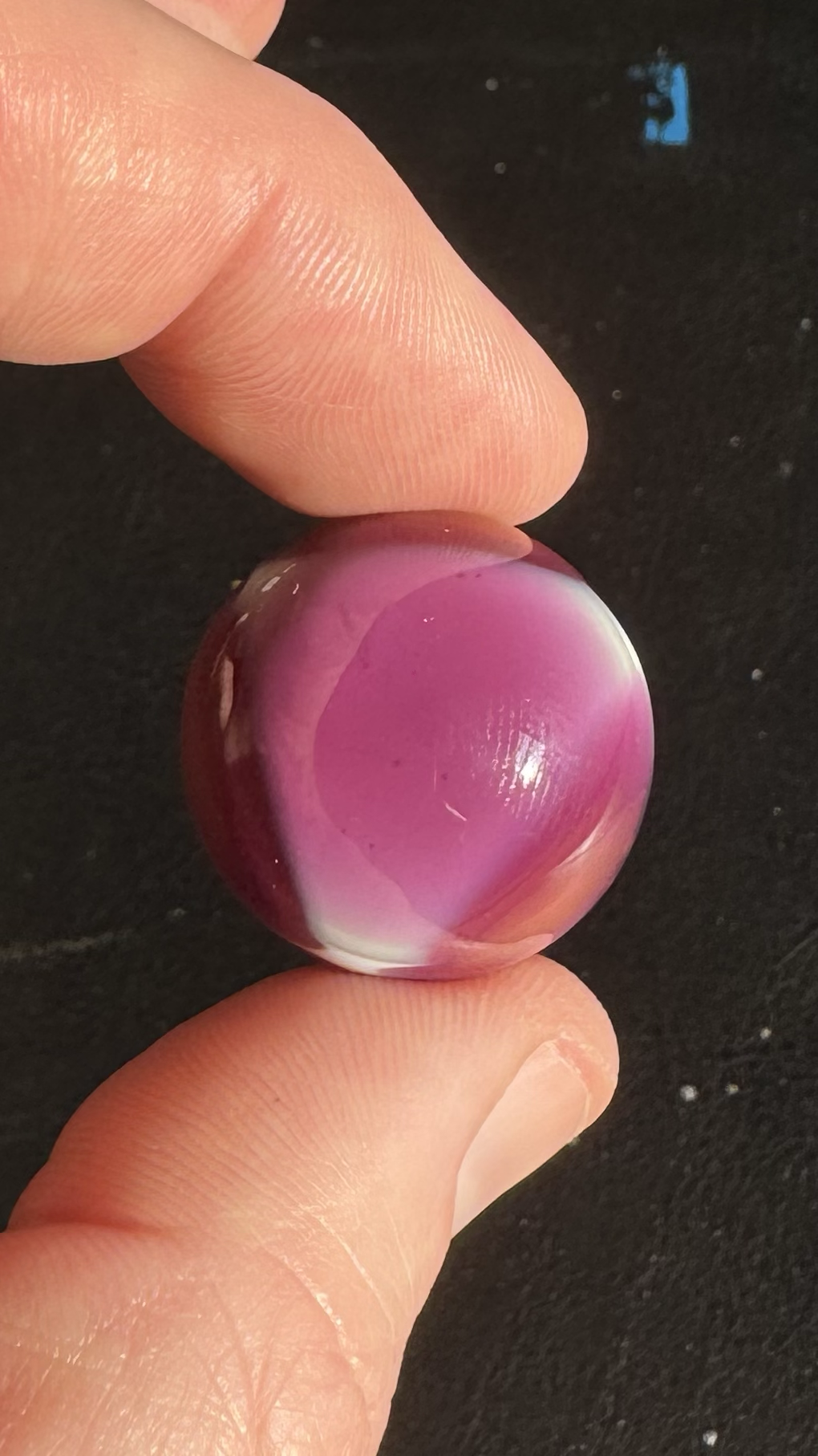A close-up of a person's fingers holding a polished marble with pink and white swirls against a black background.