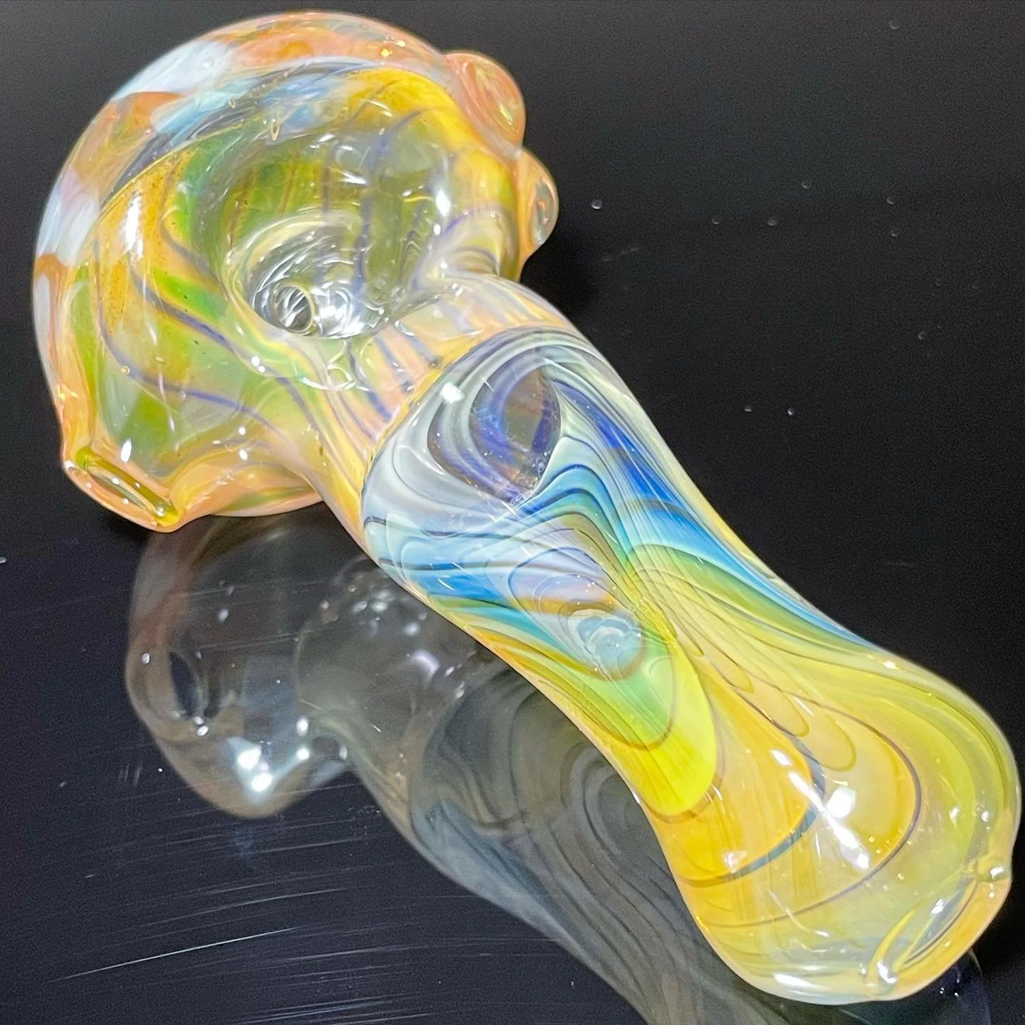 Colorful, swirled glass pipe with yellow, blue, and green accents on a reflective black surface.