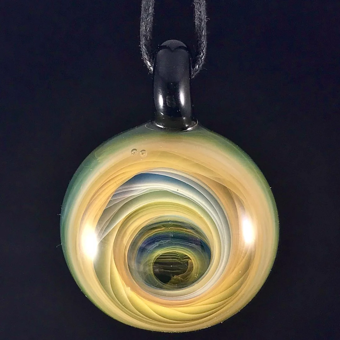 A circular glass pendant with swirling colors of yellow, green, blue, and black, hanging from a black cord against a black background.