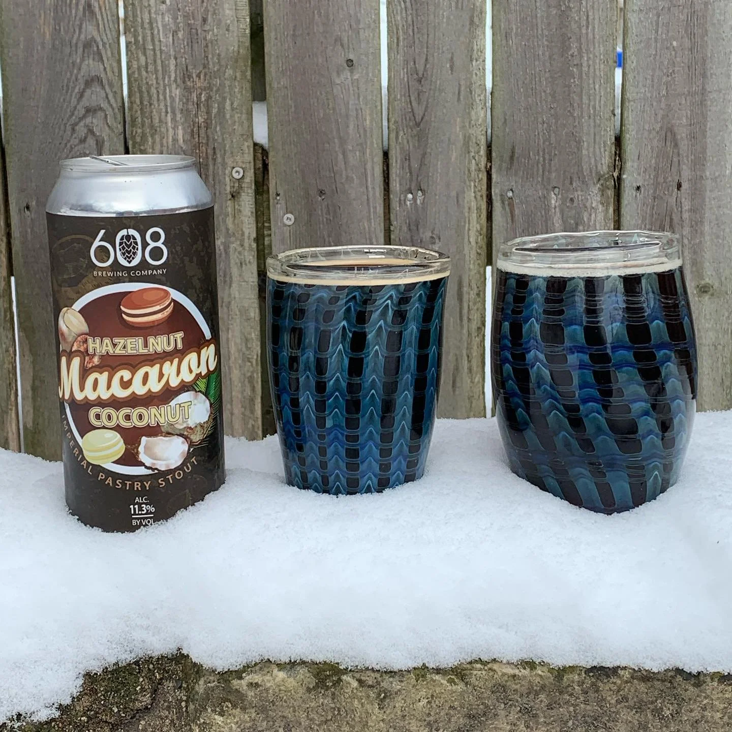 A can of 608 Brewing Company hazelnut macaroon coconut pastry stout and two glasses filled with a dark blue beverage, set on snow in front of a wooden fence.