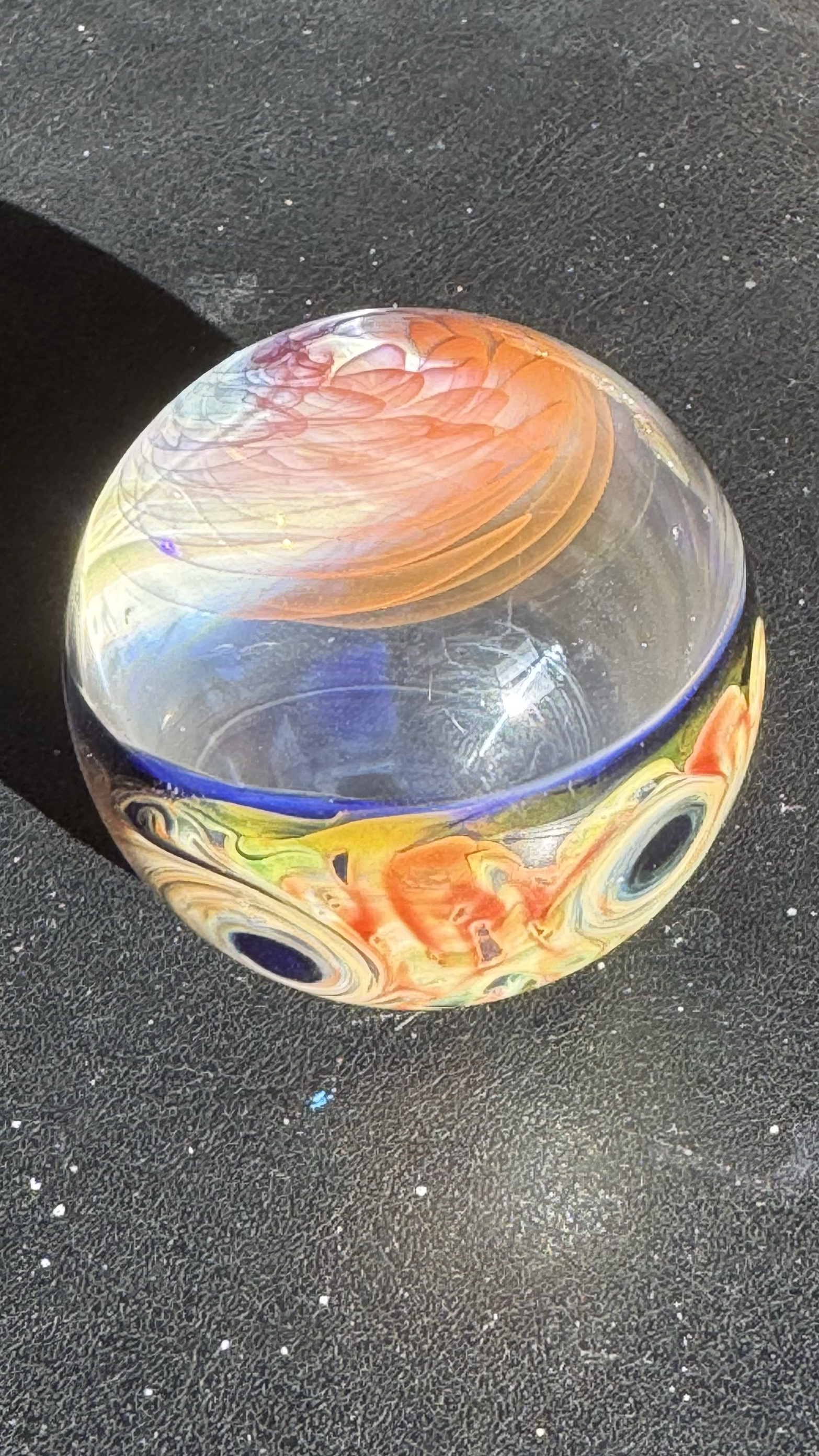 Colorful marble with orange, yellow, blue, and clear swirls resting on a textured black surface.