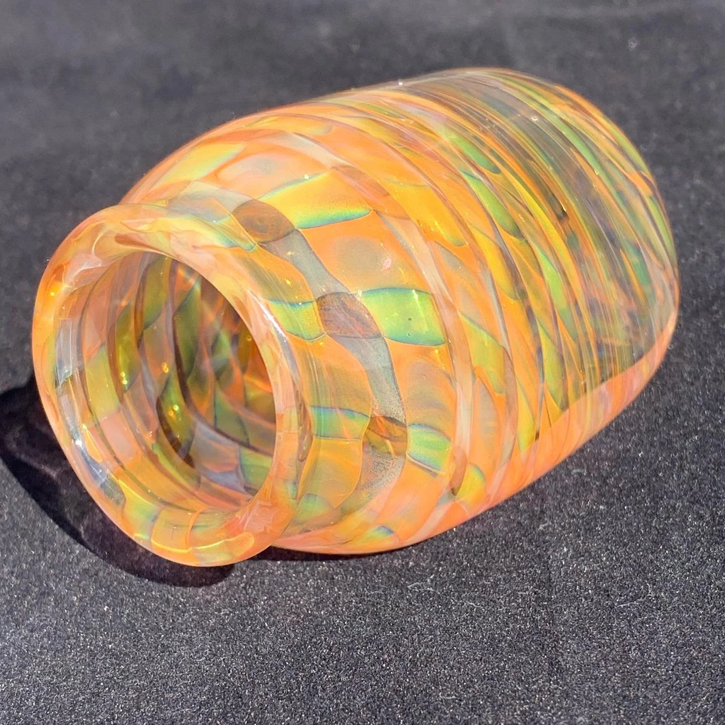 A colorful, iridescent glass mosaic cylinder with a swirling pattern, lying on a dark surface.
