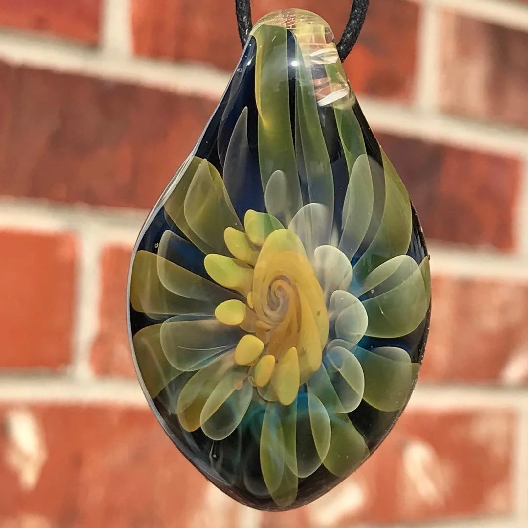 A teardrop-shaped glass pendant with a swirling yellow and green floral pattern inside, hanging against a brick wall background.