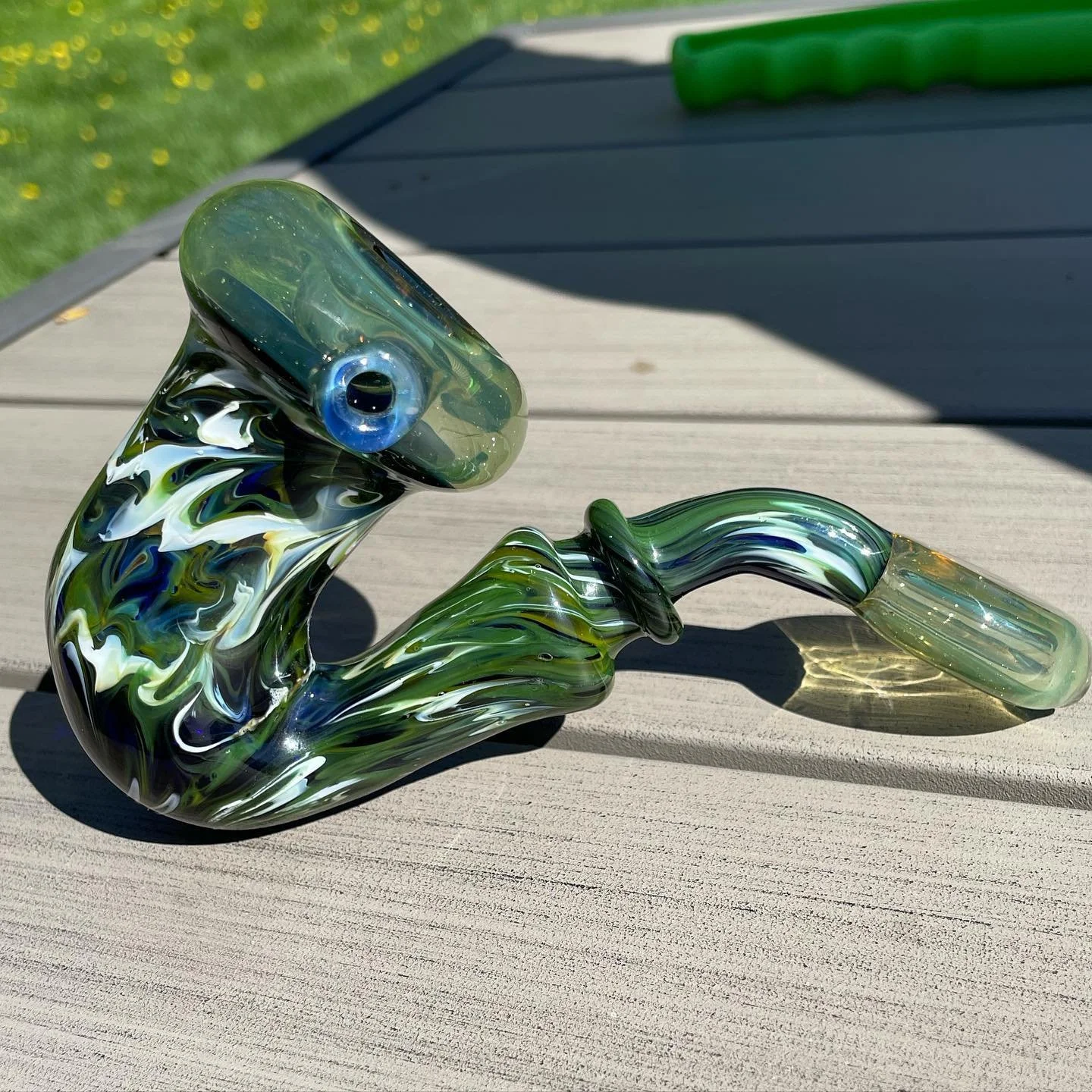 Colorful glass pipe with green, white, yellow, and blue swirls, placed on a wooden surface outdoors with grass in the background.