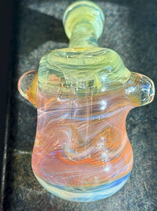 Colorful glass art piece with swirled patterns in yellow, orange, pink, and blue hues, placed on a dark surface.