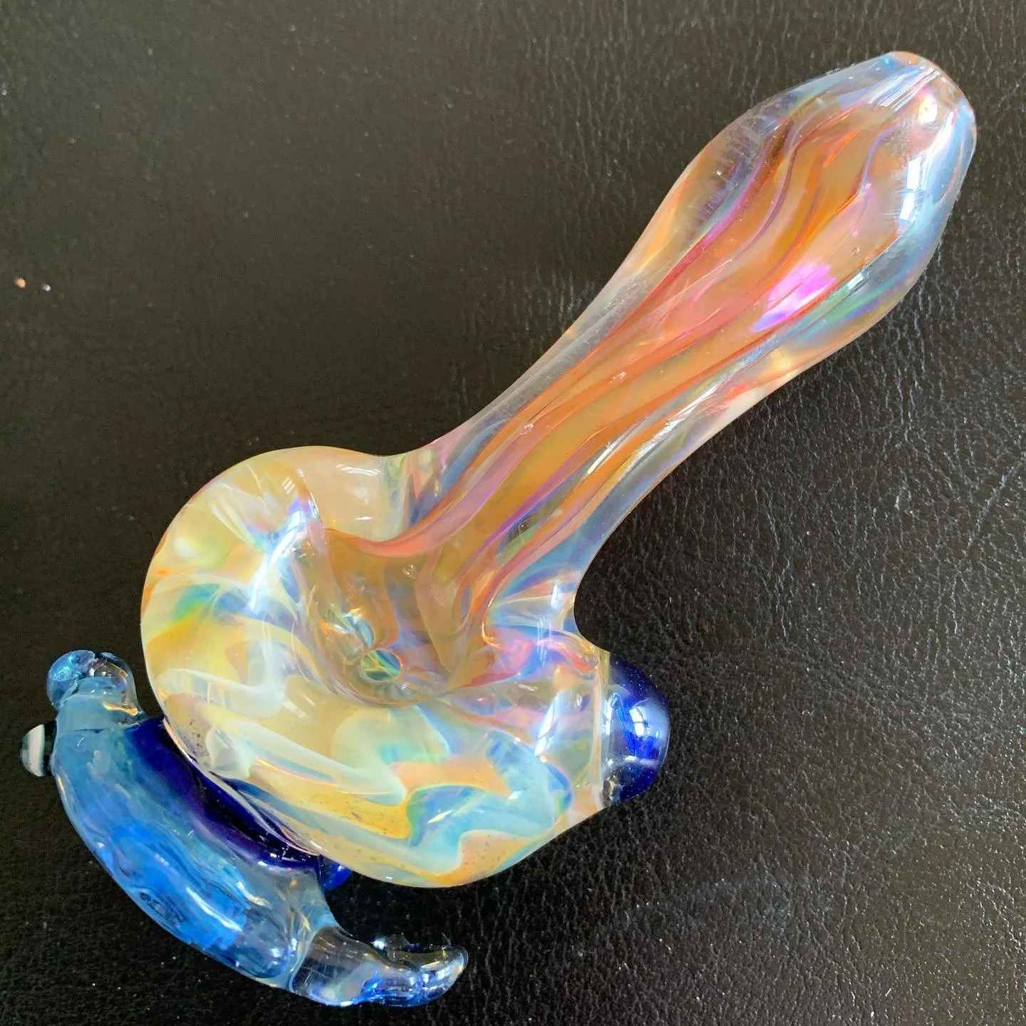 Colorful glass sculpture with a curved, elongated shape, featuring iridescent hues of yellow, pink, blue, and orange on a black background.