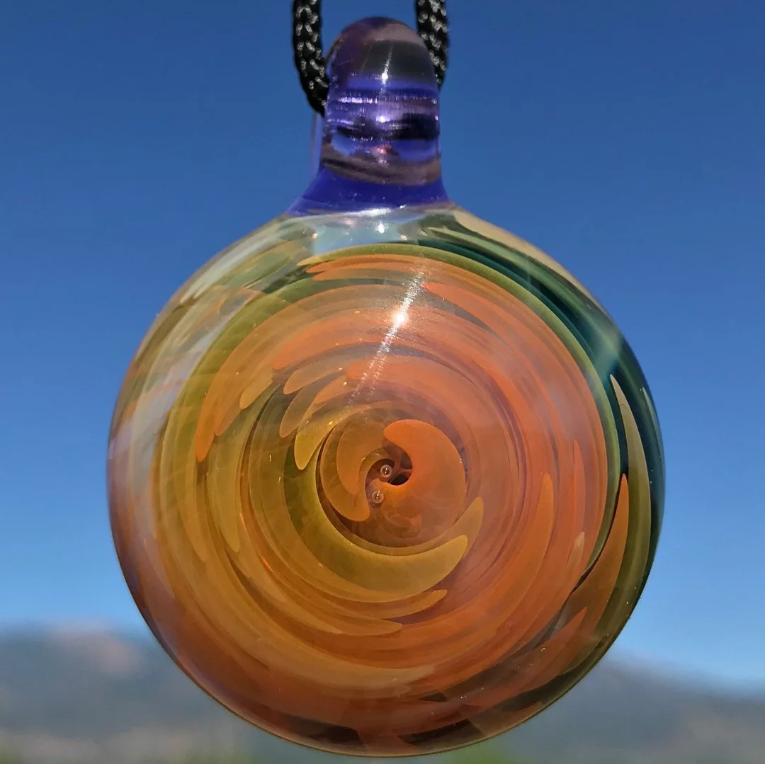 A glass pendant with a spiral swirl pattern in orange, yellow, and green, hanging against a clear blue sky.