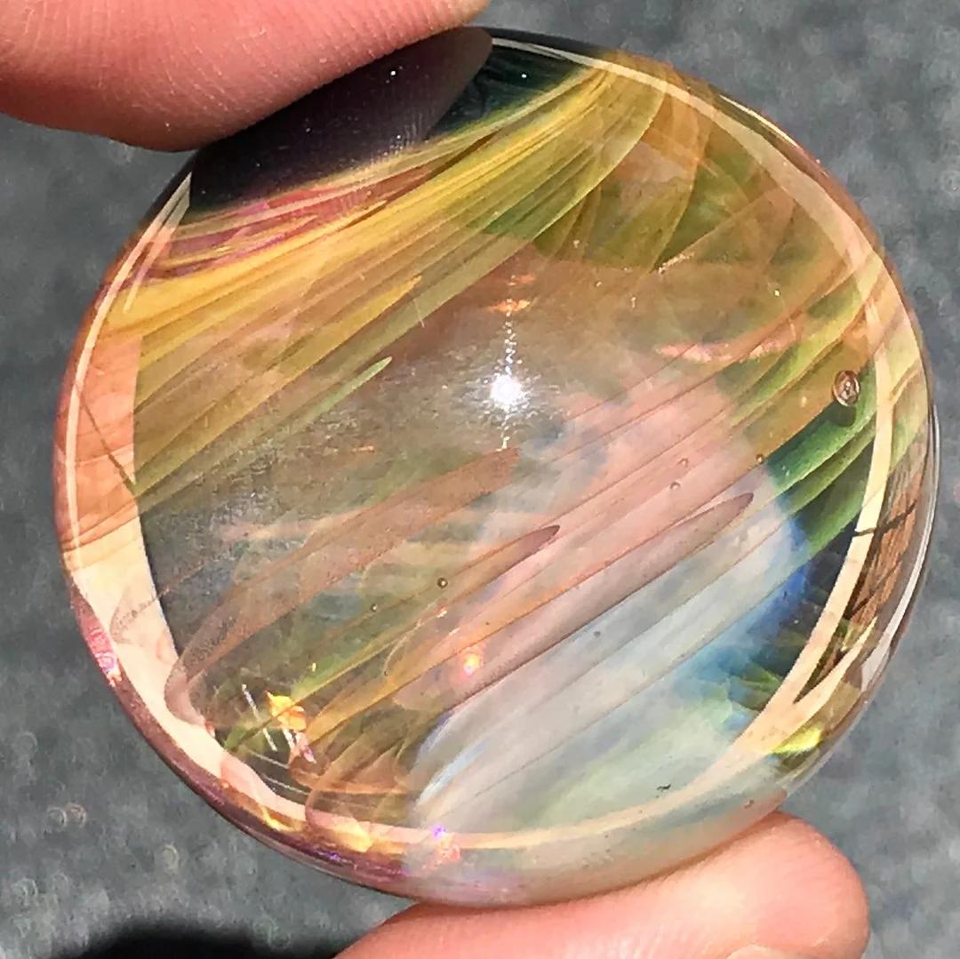 Close-up of a person's finger holding a polished, colorful, oval-shaped gemstone or glass sphere with swirling patterns in green, pink, yellow, and black, against a gray background.
