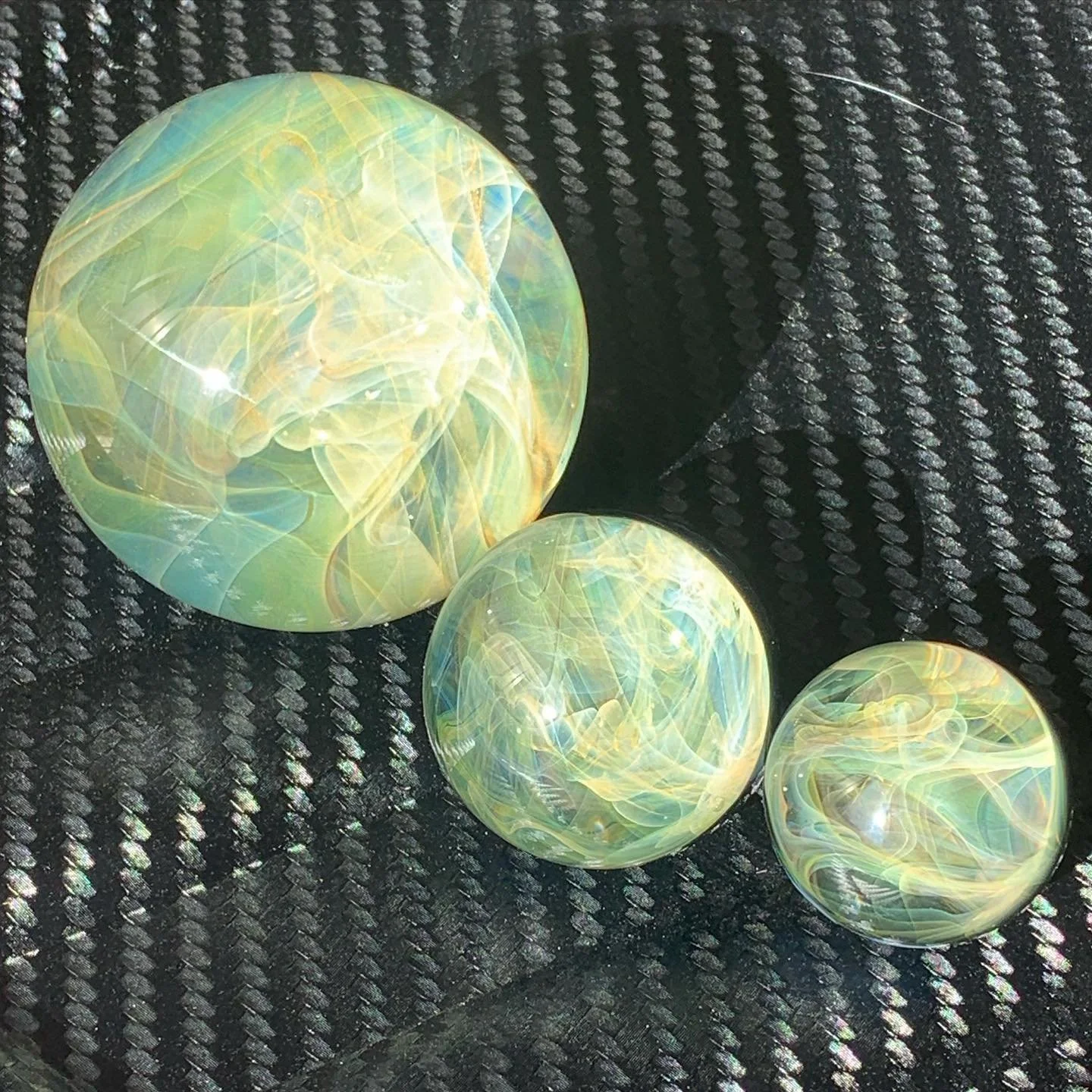 Three glass marbles with gold and silver fume chaos swirling patterns on a black textured surface.