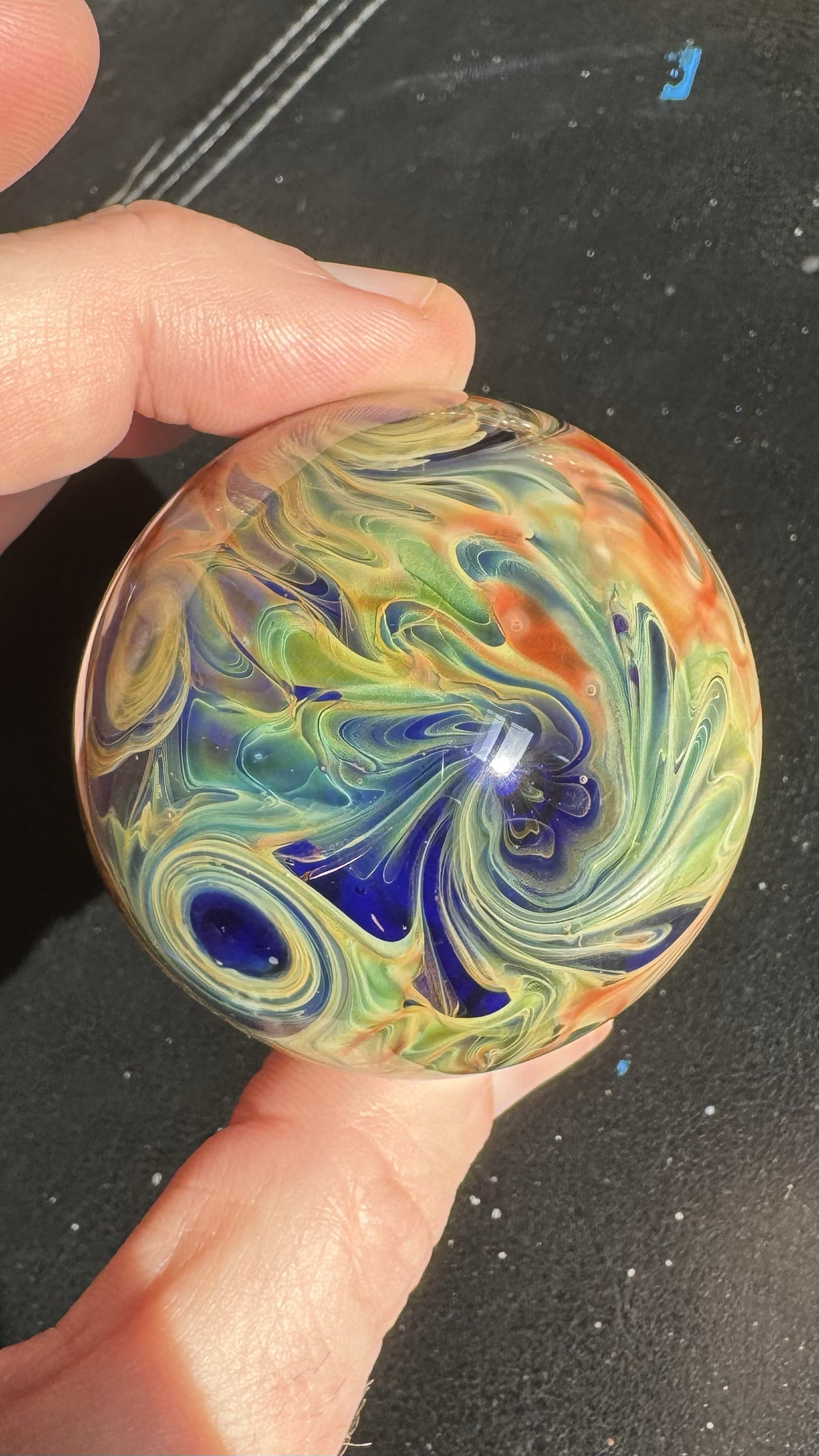 Hand holding a colorful, swirled glass marble over a black surface with speckles.