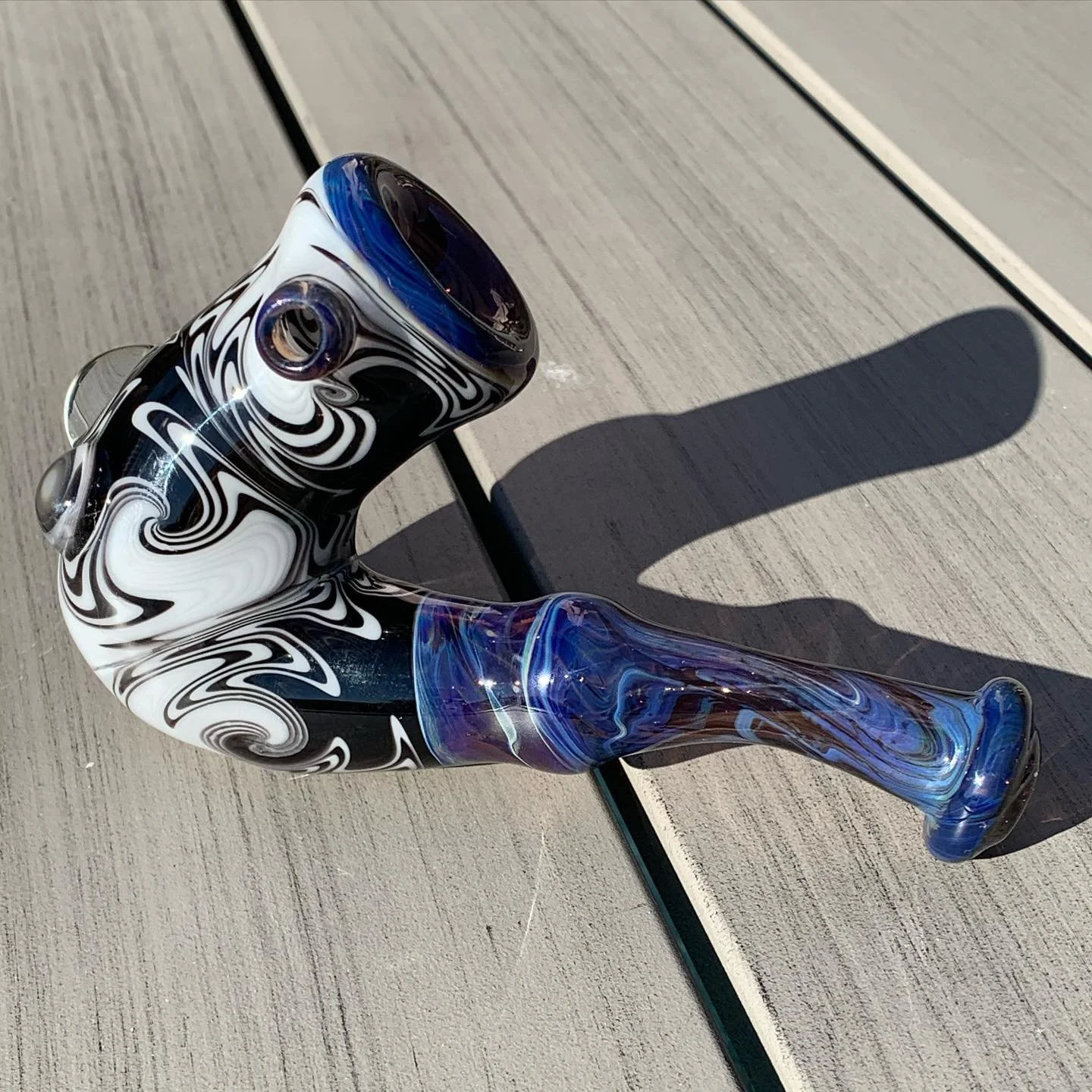 A colorful glass pipe with black, white, blue, and purple swirls resting on a wooden surface, casting a shadow.