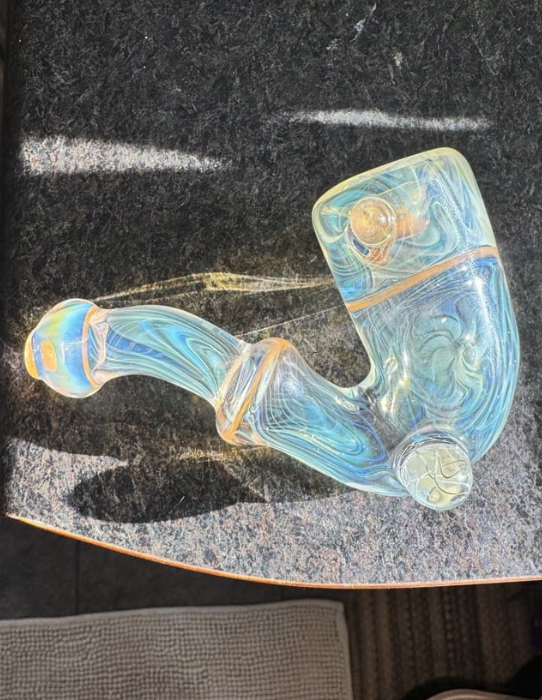 A glass pipe with swirled blue, yellow, and clear colors, resting on a dark textured surface.