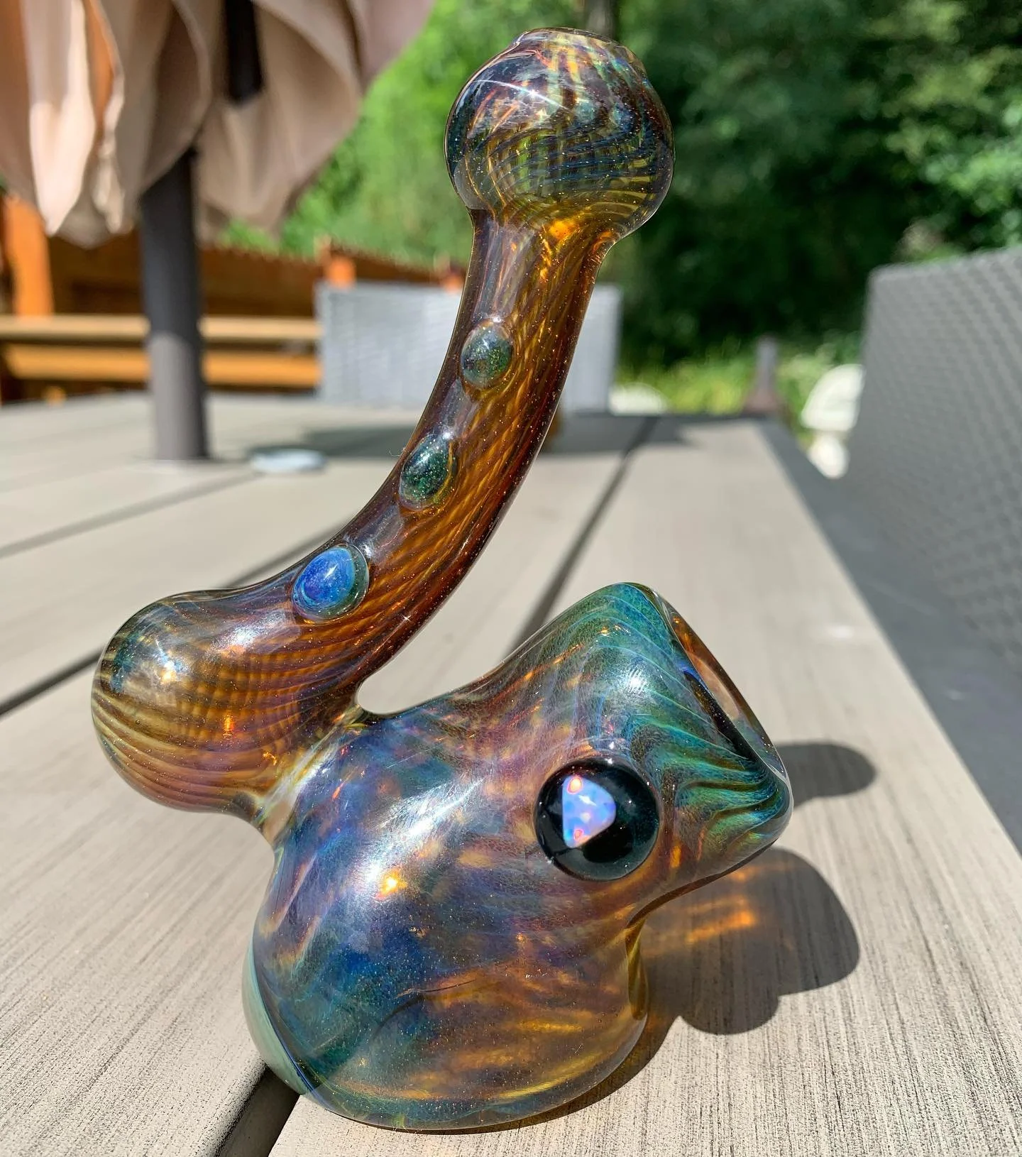 Colorful glass sculpture resembling a rabbit with a curved, elongated neck and large black eye, placed on a wooden outdoor table with green trees in the background.