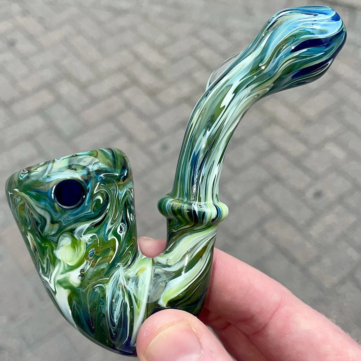 Close-up of a colorful glass pipe with swirled green, blue, white, and black patterns, held in a person's hand against a gray paved background.