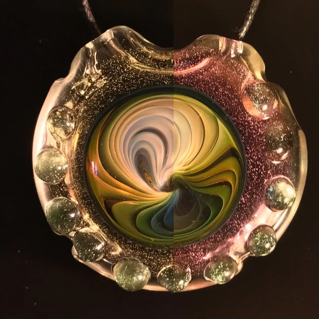 A colorful glass art piece with a swirling pattern in the center, surrounded by a clear, wavy glass border with bubble-like details against a black background.