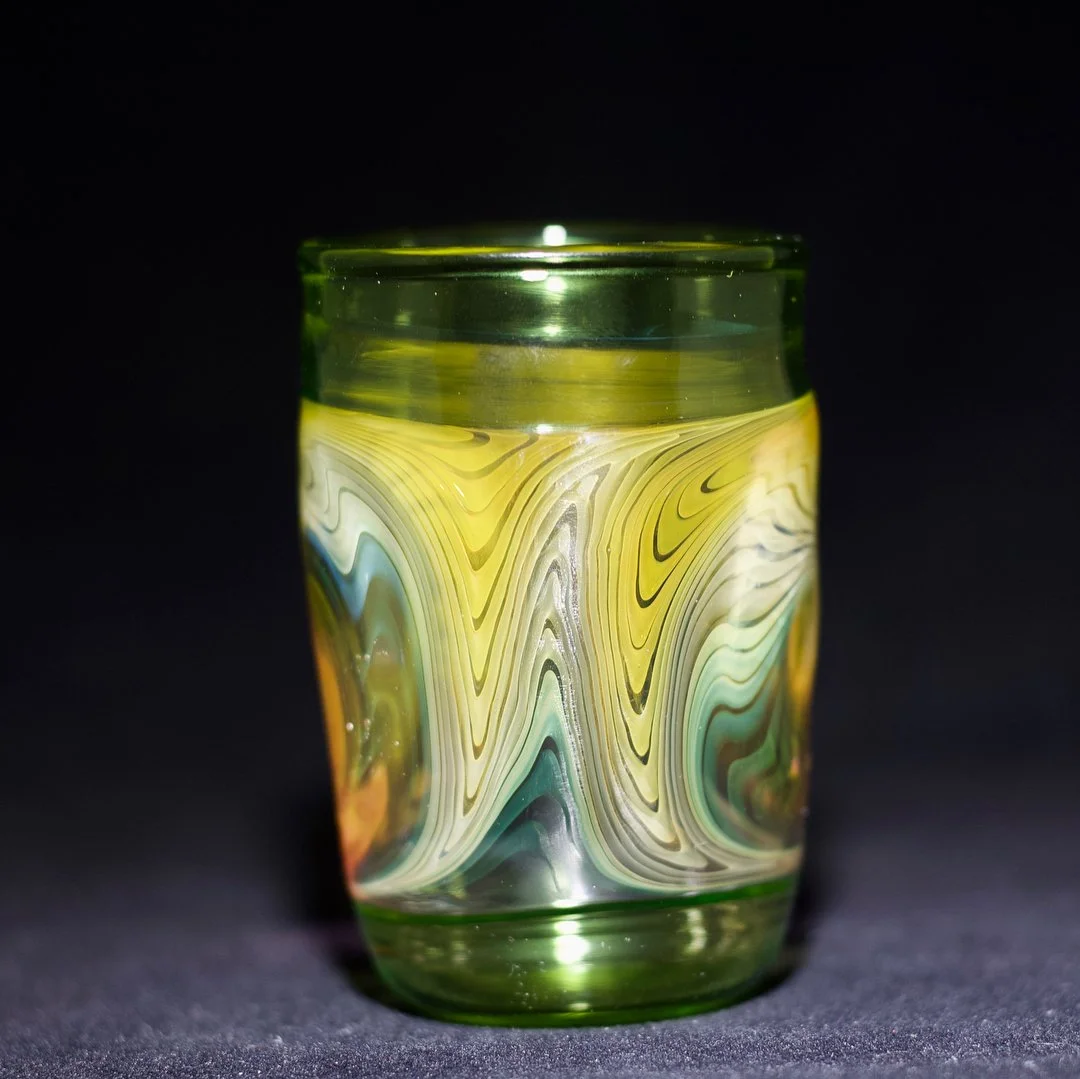 A colorful, marbled glass tumbler with yellow, white, green, and orange swirling patterns, set against a black background.