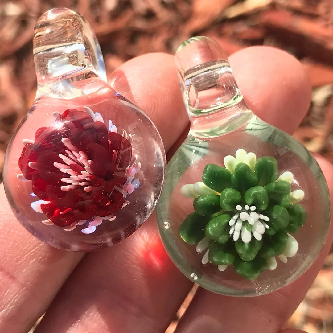 Two glass bottle pendants, one with a red and white flower and the other with a green and white flower, held against a background of dry leaves