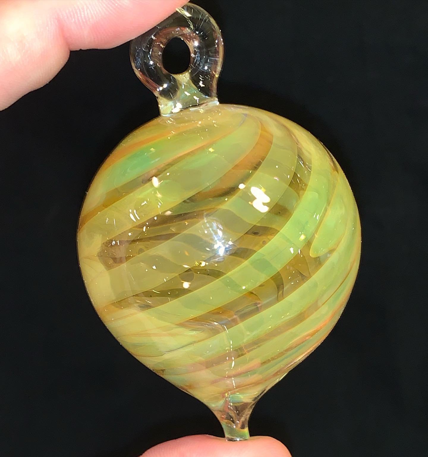 Hand holding a yellow and orange striped glass ornament with a loop on top, against a black background.