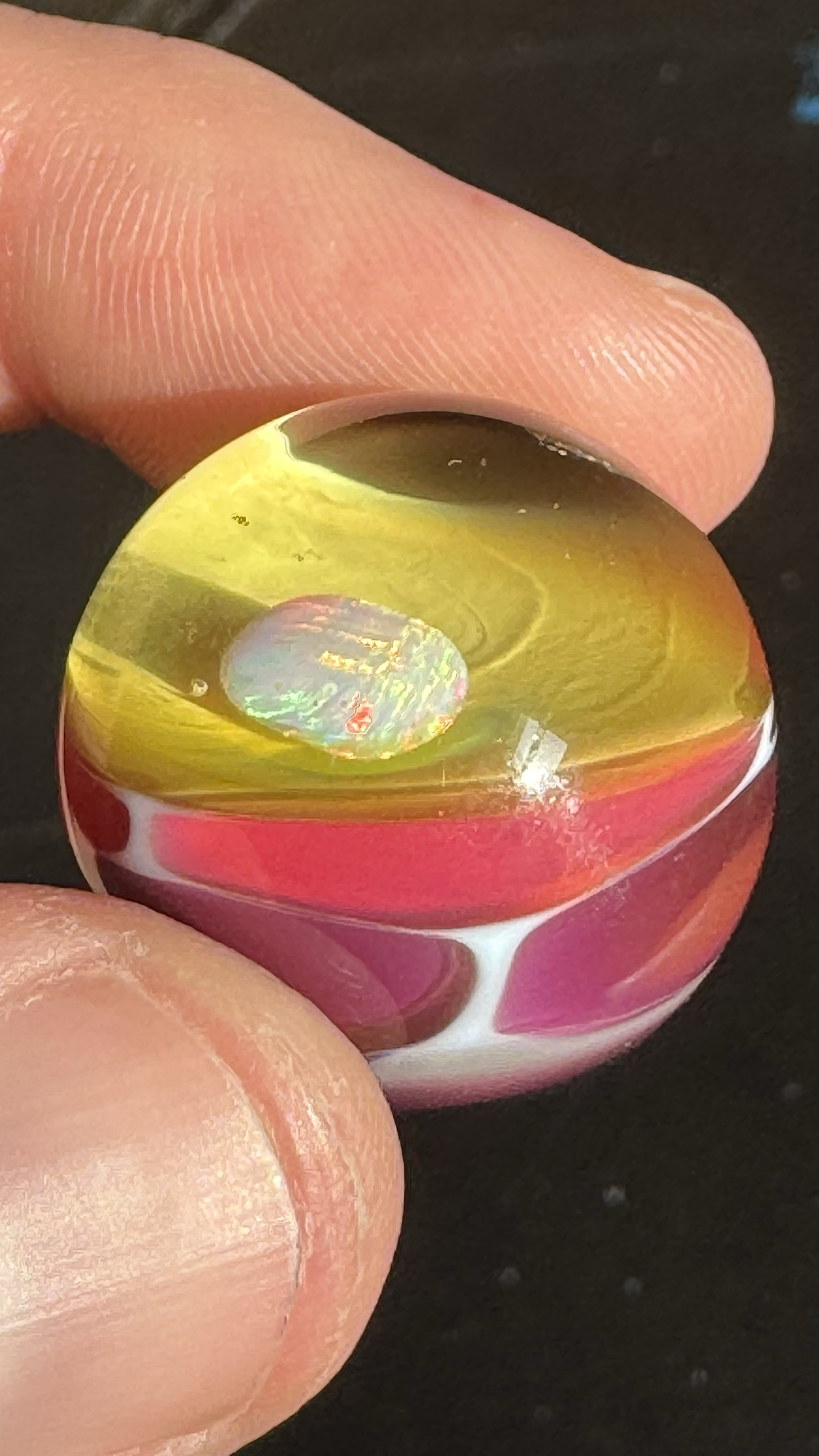 Close-up of a person's fingers holding a small, colorful glass marble with yellow, pink, and iridescent patterns.