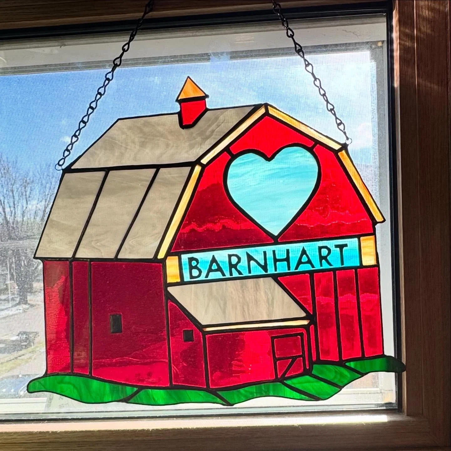 Colorful stained glass window depicting a red barn with a heart-shaped window, a blue sign reading 'BARNHART', green grass at the bottom, and a background of a cloudy sky seen through the window.