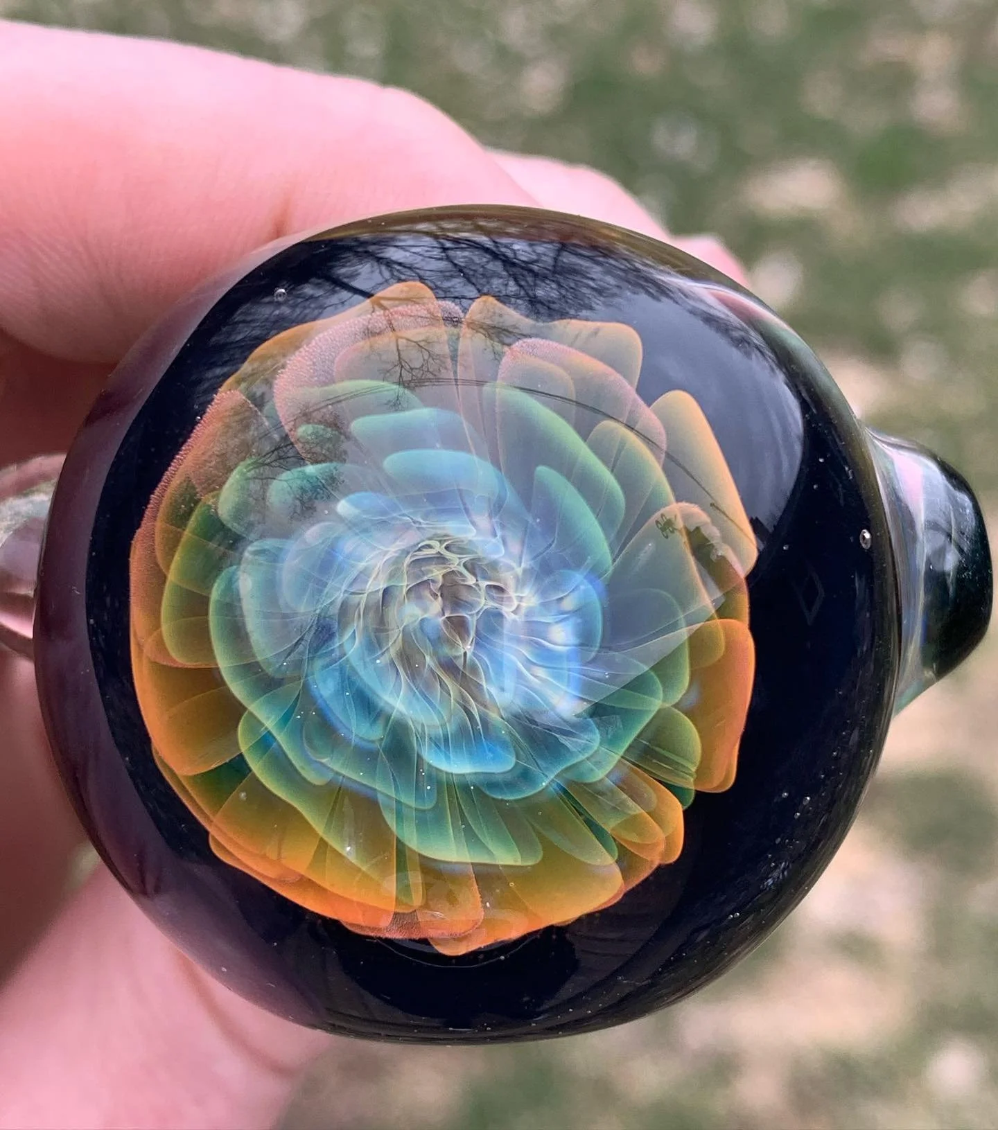Close-up of a glass paperweight with colorful swirling patterns inside, held by a person's hand outdoors.