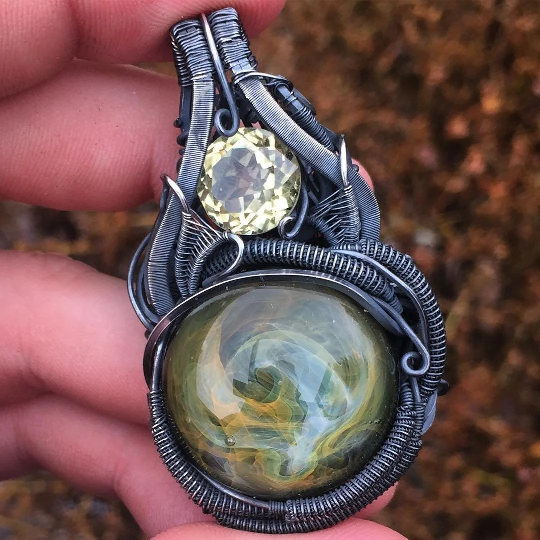 A handcrafted wire-wrapped pendant featuring a large, round, yellow glass cabochon with a swirling design, and a smaller, faceted, yellow gemstone above it, all set against a blurred outdoor background.