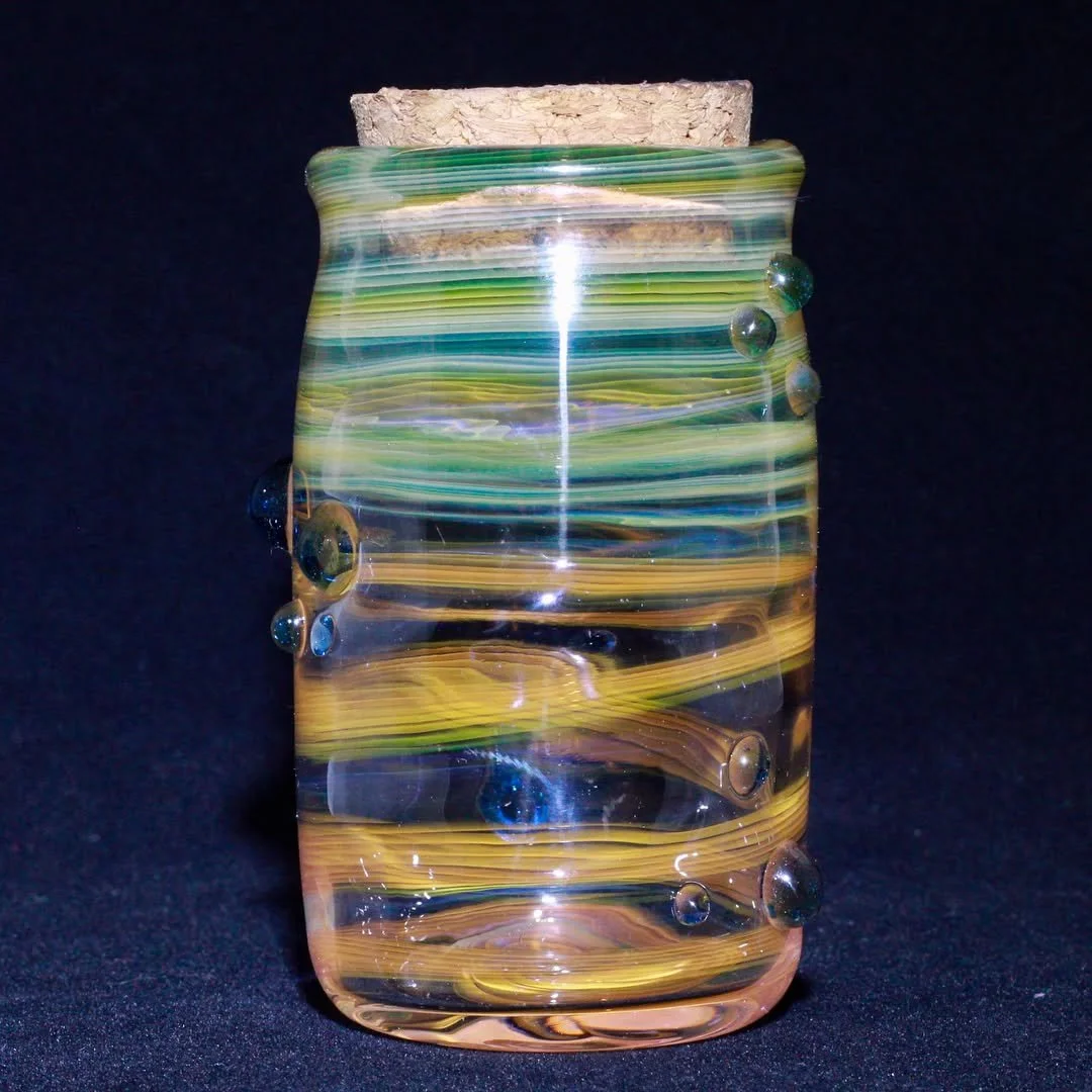 Colorful glass vase with green, blue, yellow, and purple swirls and bubbles, with a piece of cork on top, against a black background.