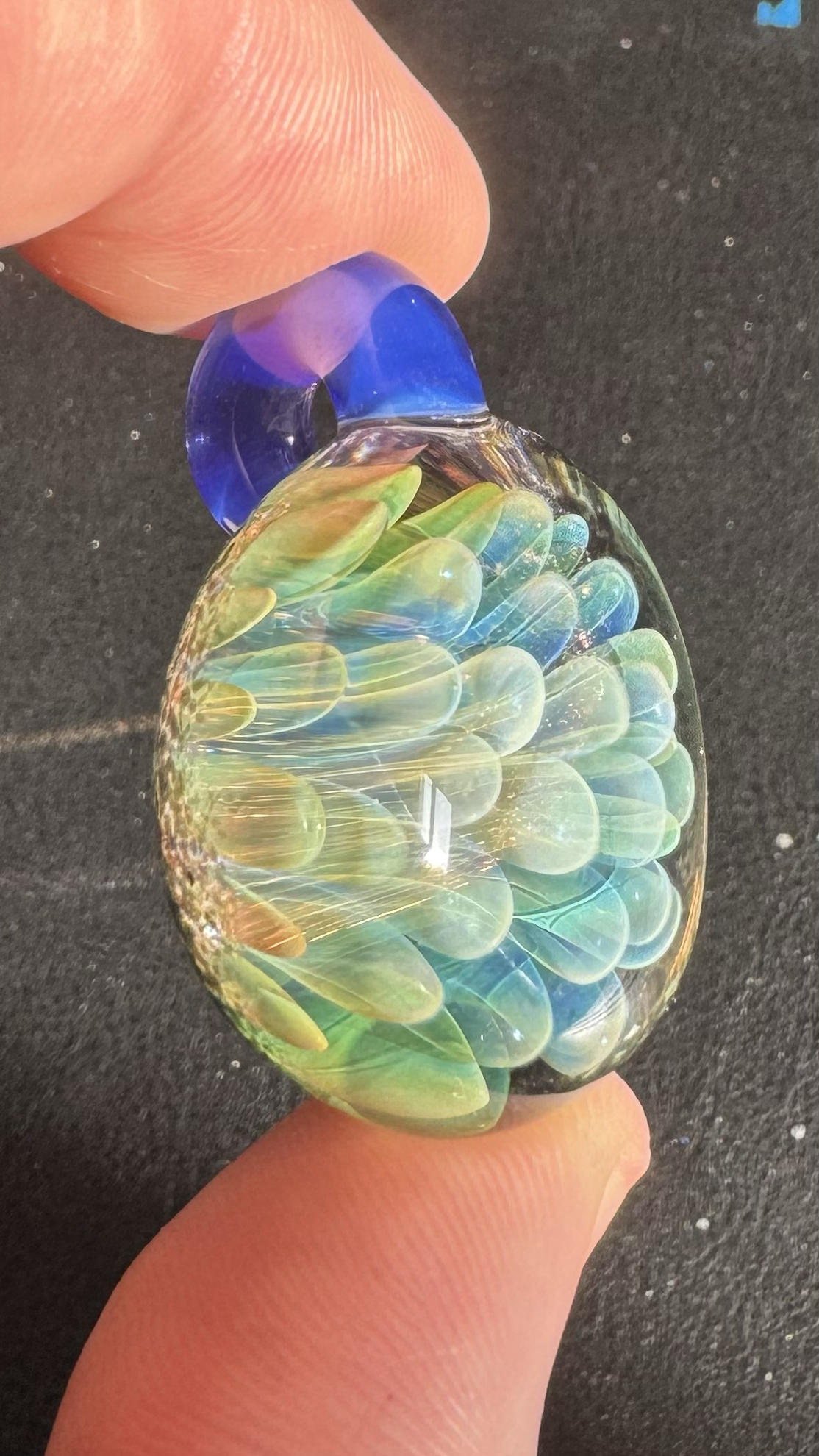 A close-up of a glass pendant with colorful, iridescent bubbles inside, held between two fingers.
