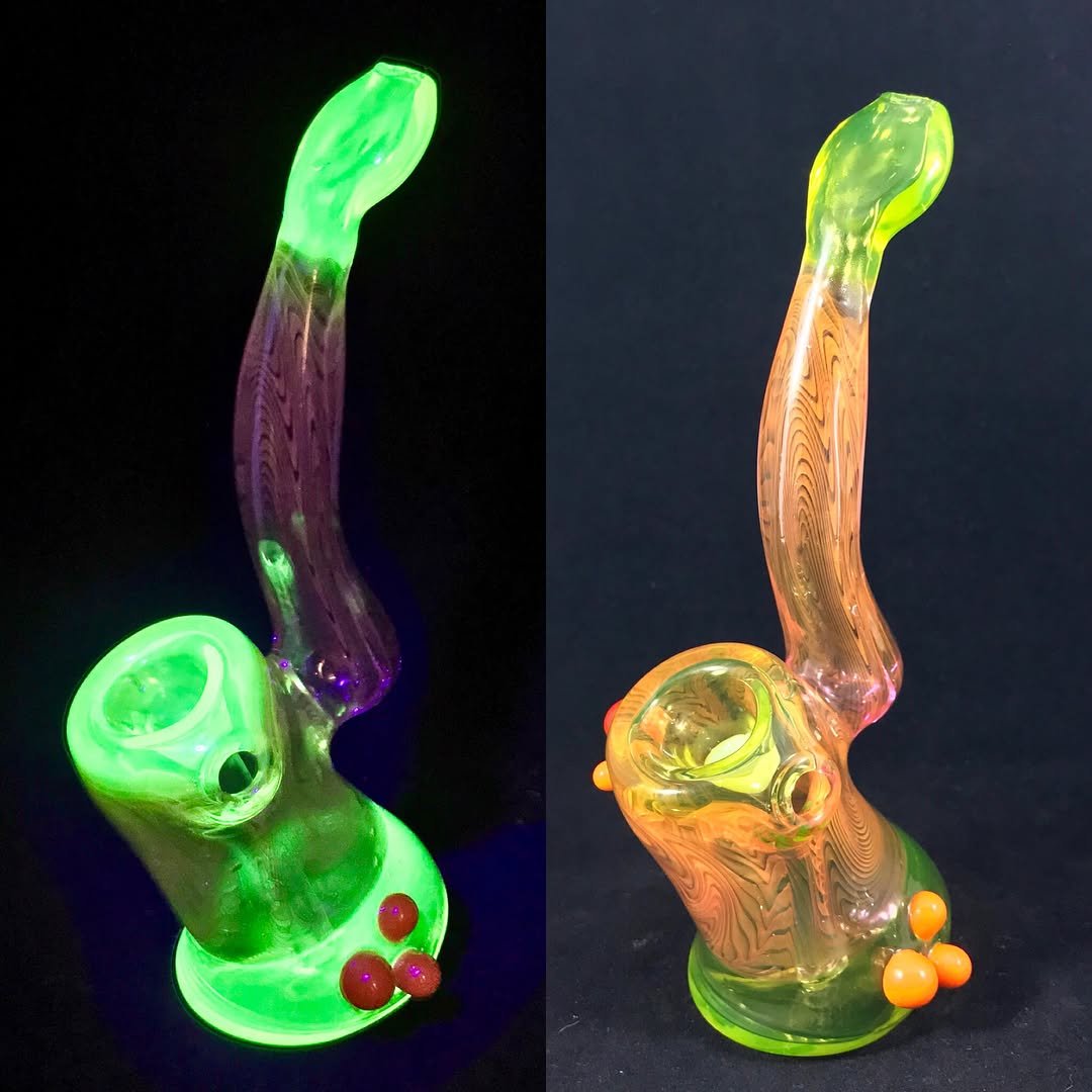 Two glass smoking pipes, one glowing green in the dark and the other with orange accents, both with a curved shape.